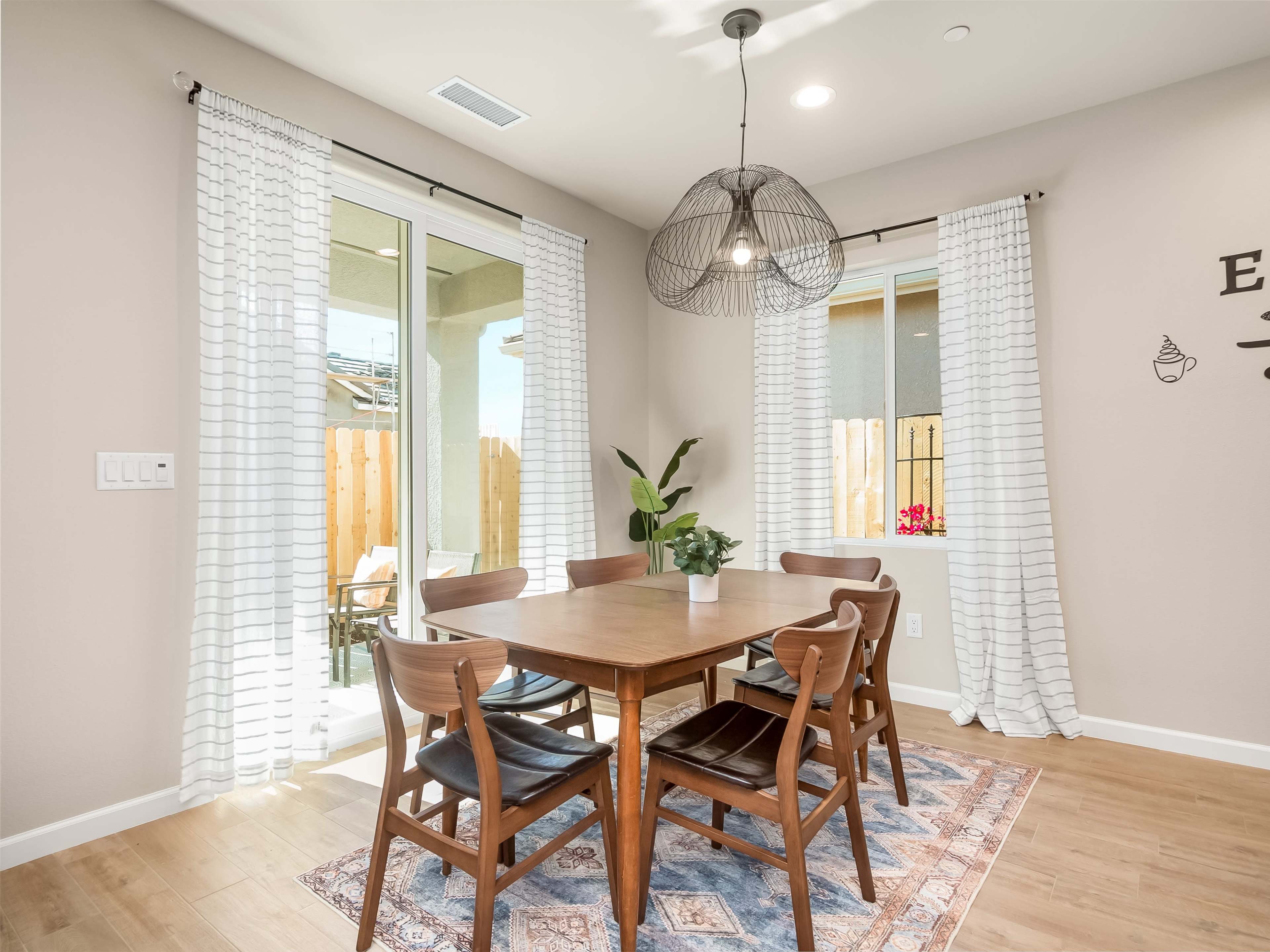 A dining area features a wooden table surrounded by six chairs, with large windows and a plant beside them.