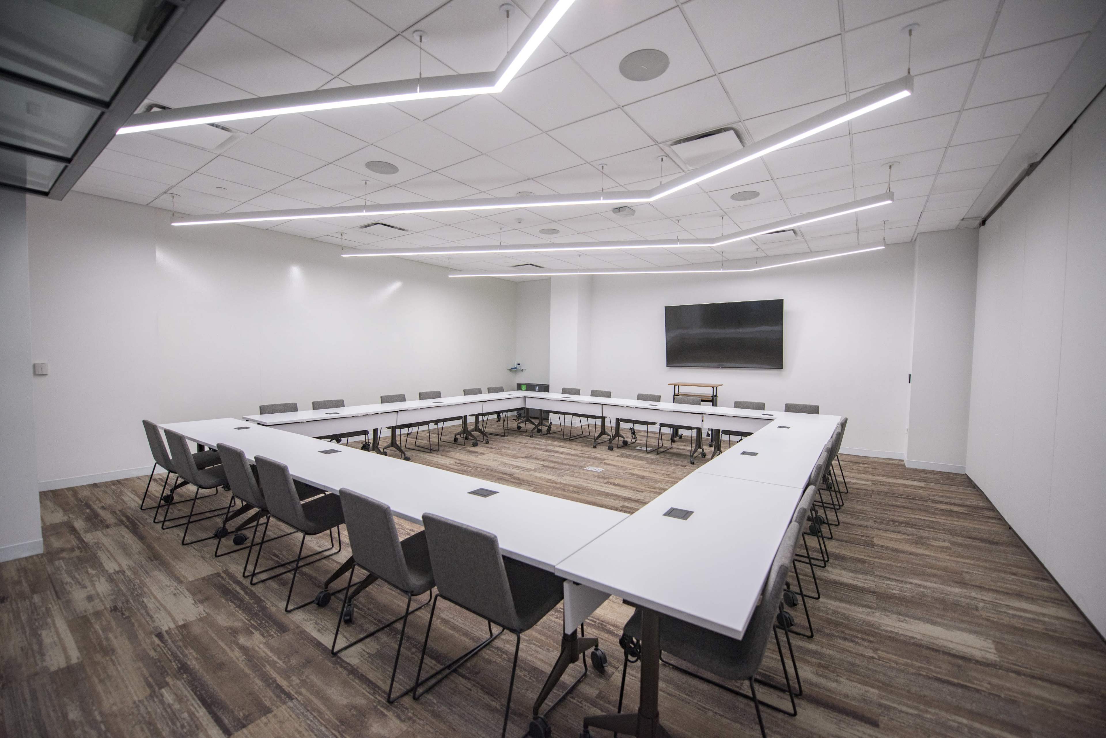 A large, modern conference room features a rectangular arrangement of white tables and gray chairs surrounding a central open space, with a television mounted on the wall.