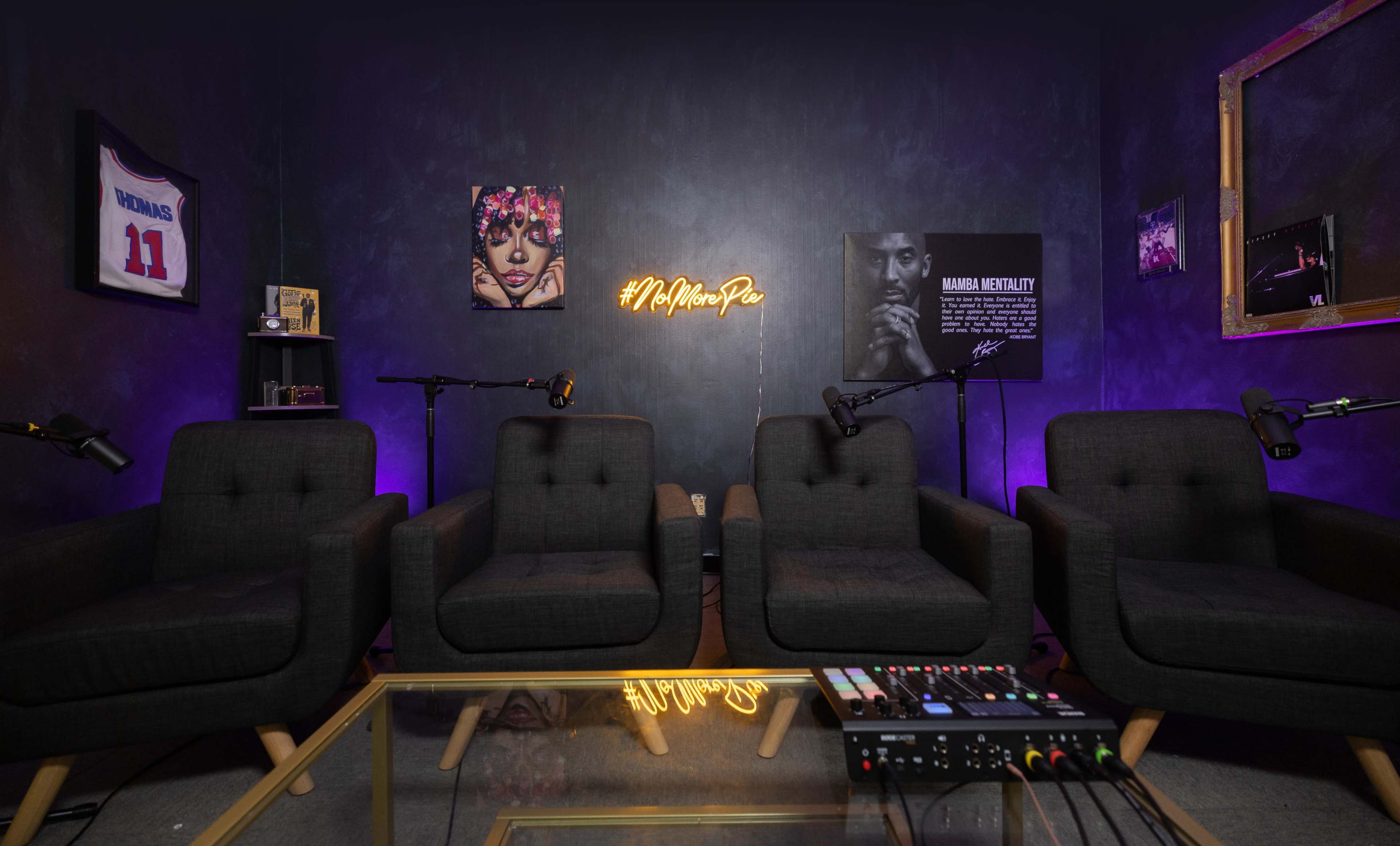 A small, modern podcast studio with four black chairs, a glass table, and two microphones, illuminated by a neon sign on the wall.