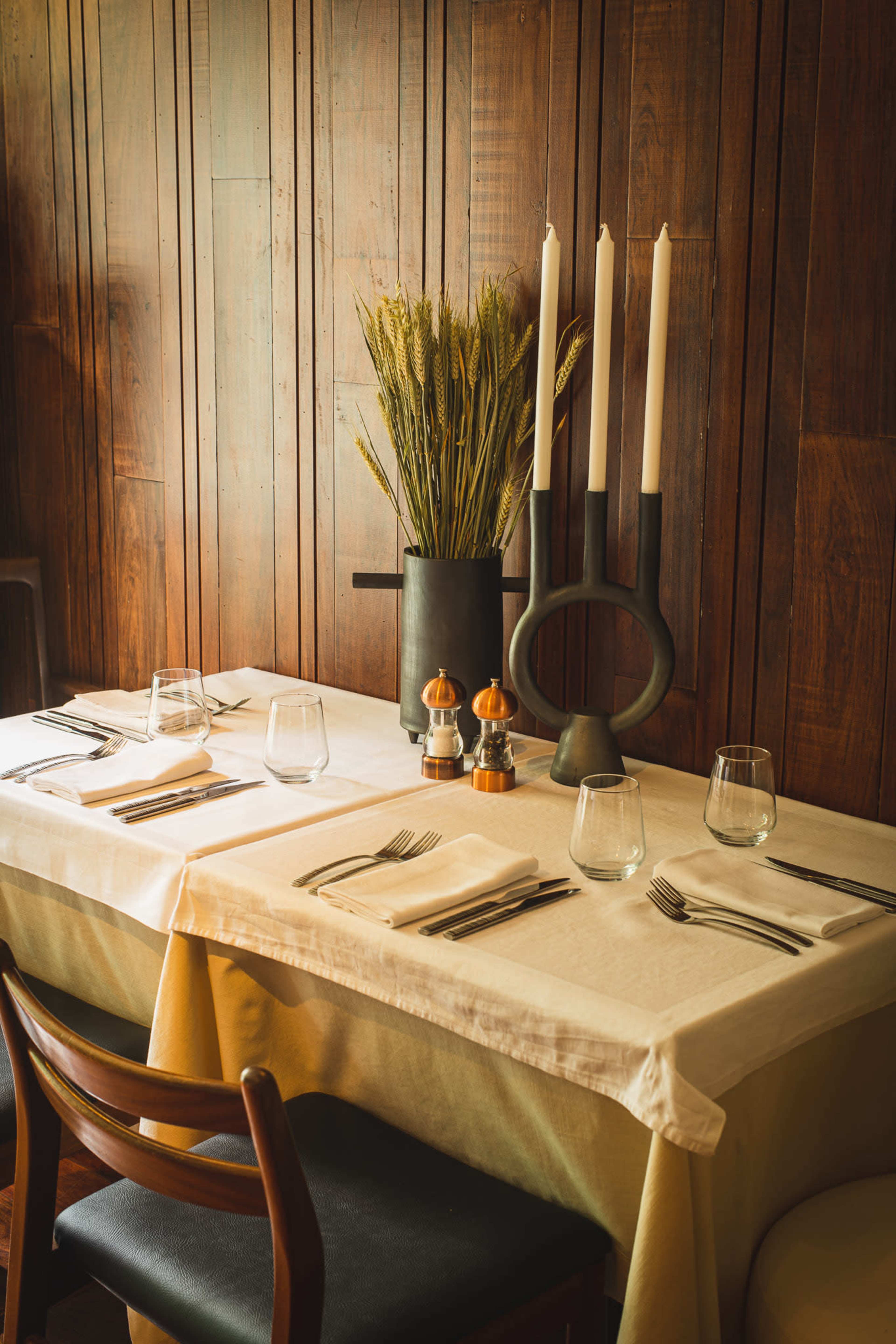 A dining table is elegantly set with glassware and cutlery, surrounded by wooden walls, and adorned with a vase of wheat and three candles.
