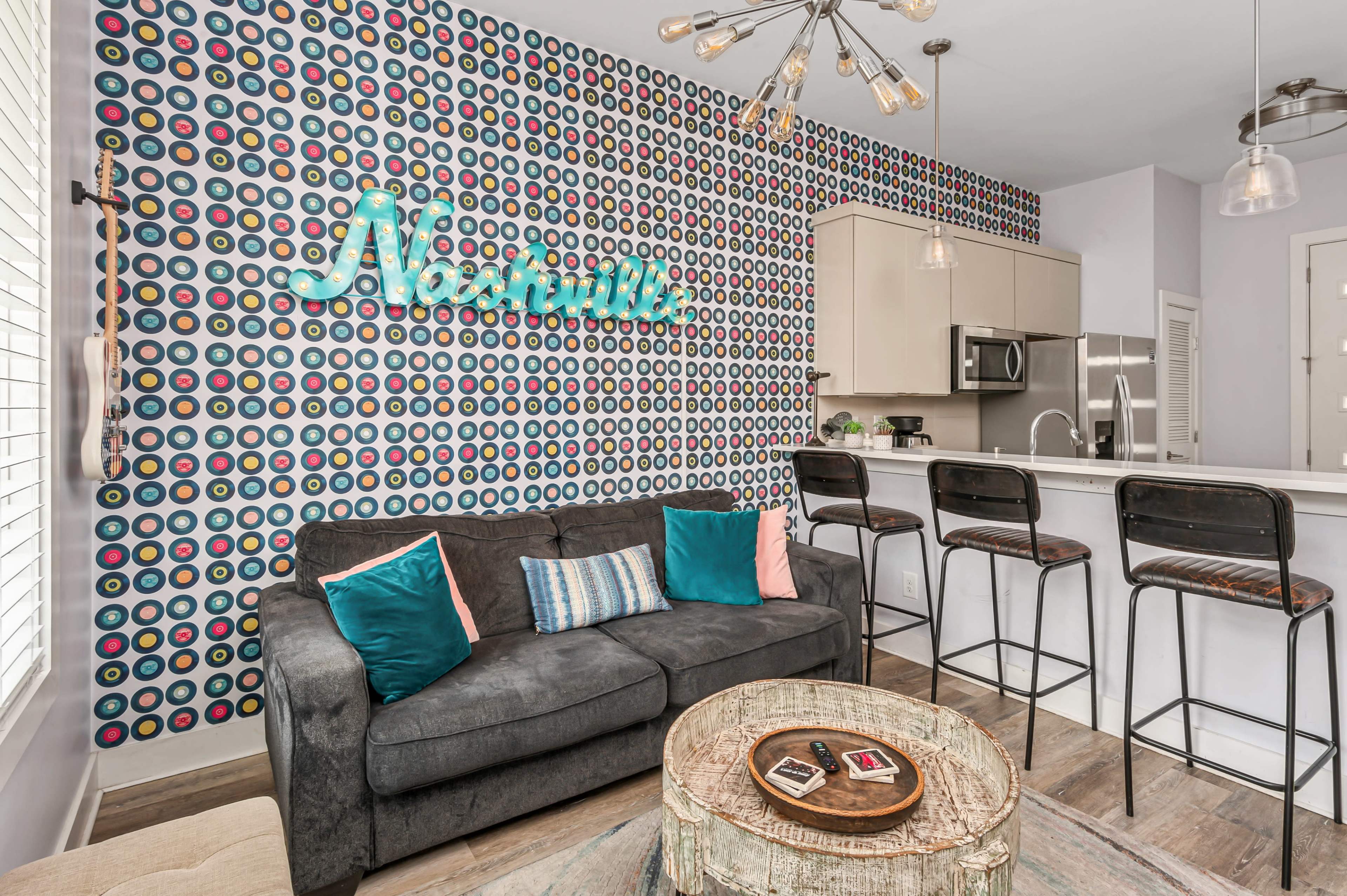 A modern living room features a patterned accent wall, a dark sofa with colorful pillows, and a bar with three stools overlooking a kitchen.