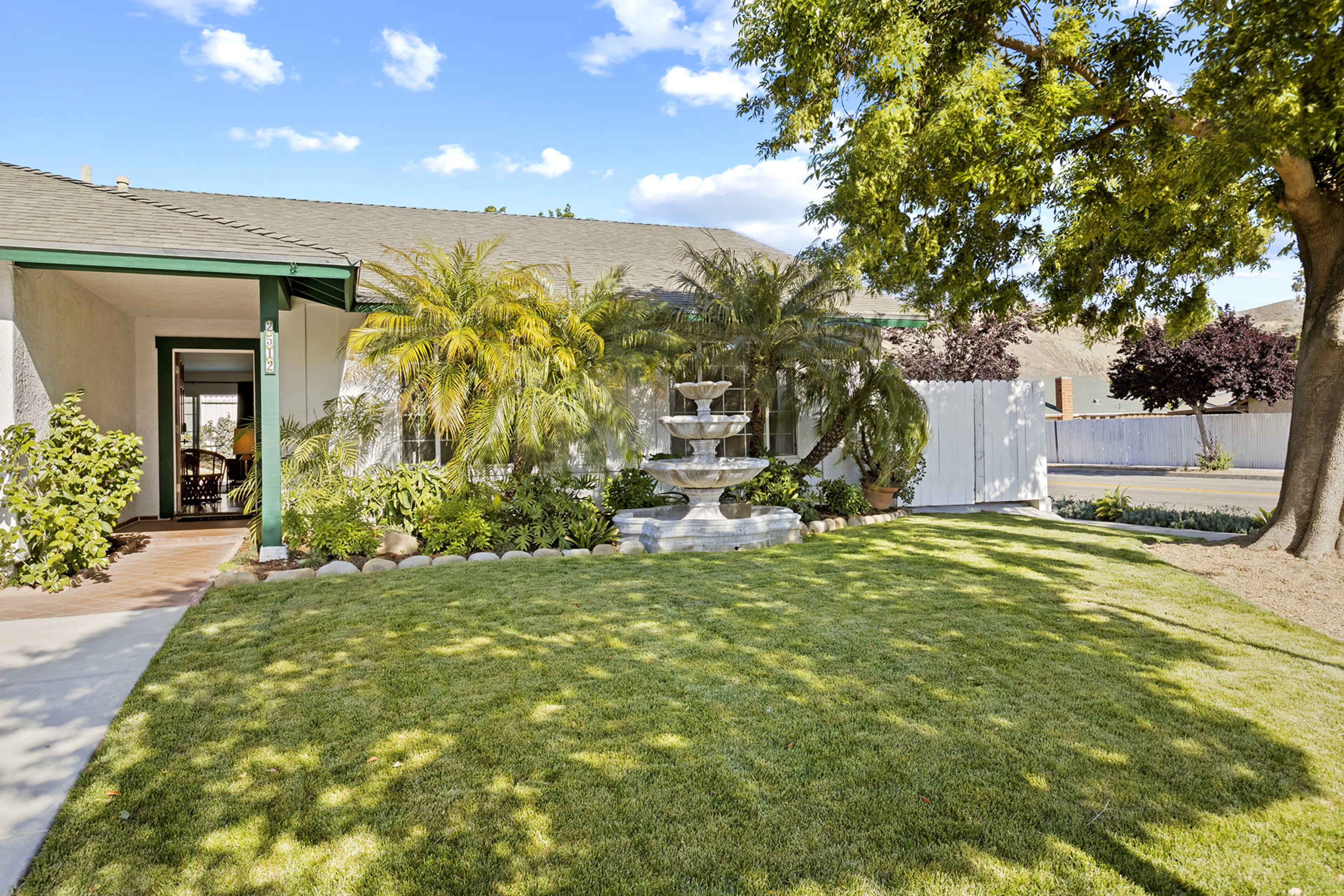 The image shows a well-maintained front yard featuring a fountain, lush greenery, and a pathway leading to a single-story house.