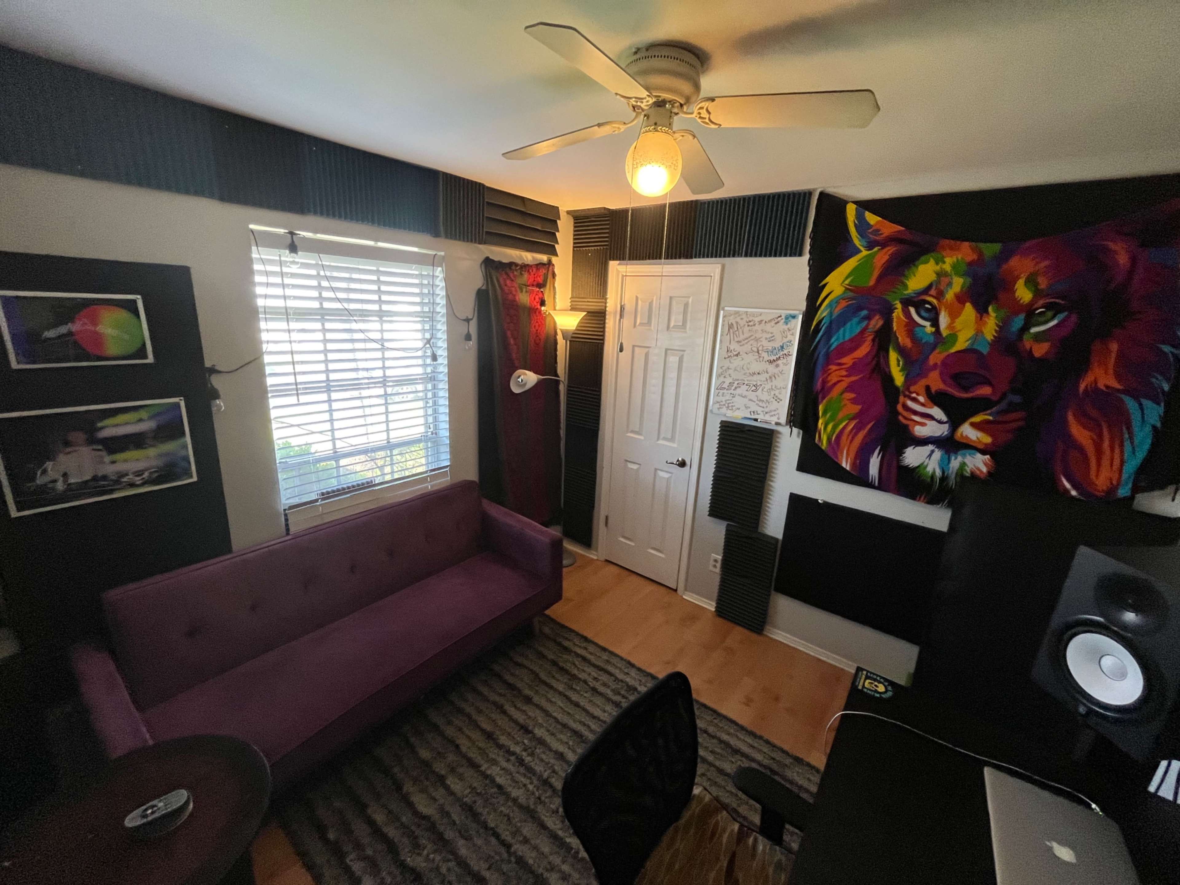 The image shows a well-decorated room featuring a purple couch, a lion-themed tapestry on the wall, a desk with audio equipment, and soundproofing panels.
