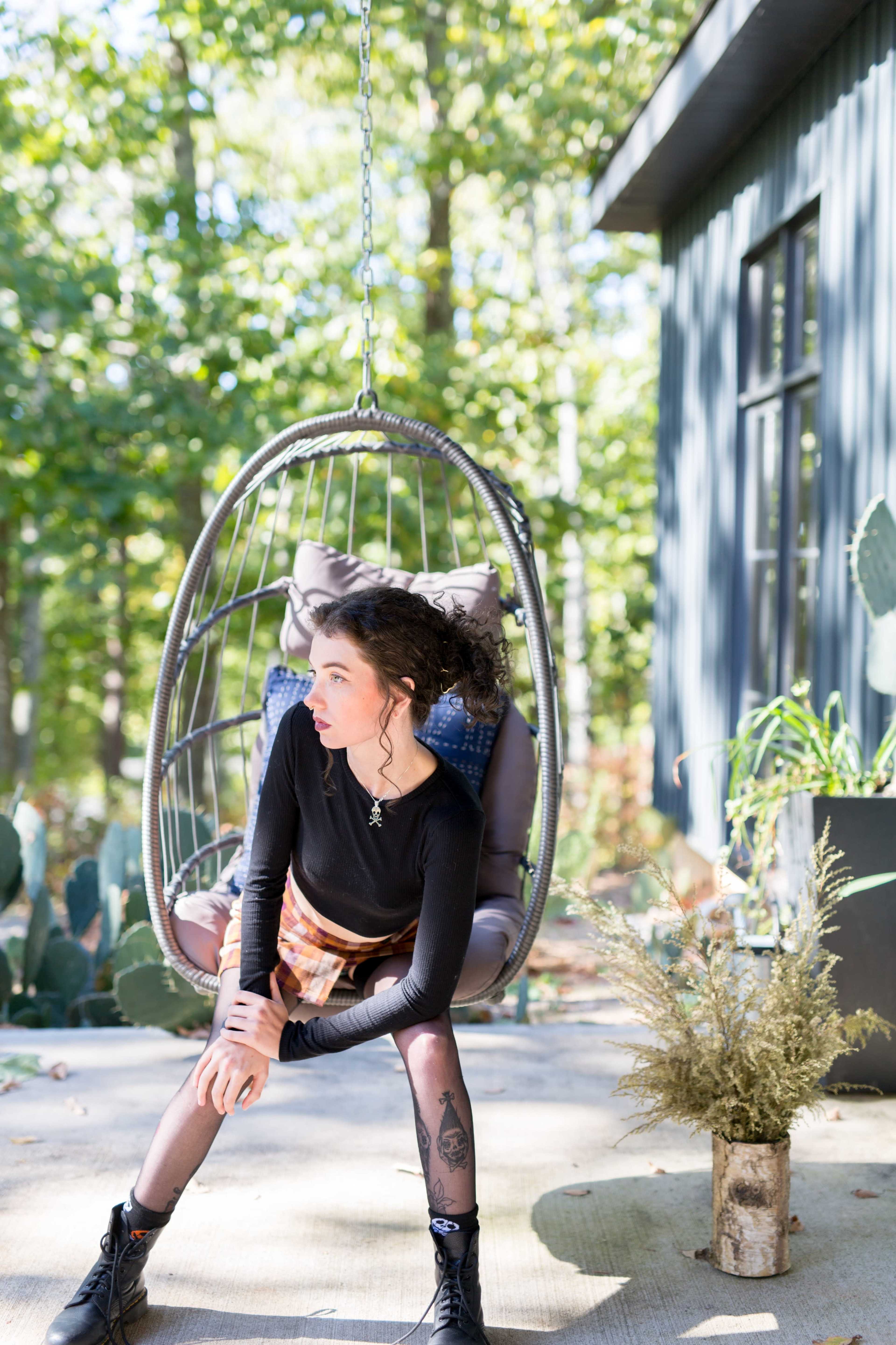 A woman sits in a hanging chair on a patio surrounded by greenery and potted plants.