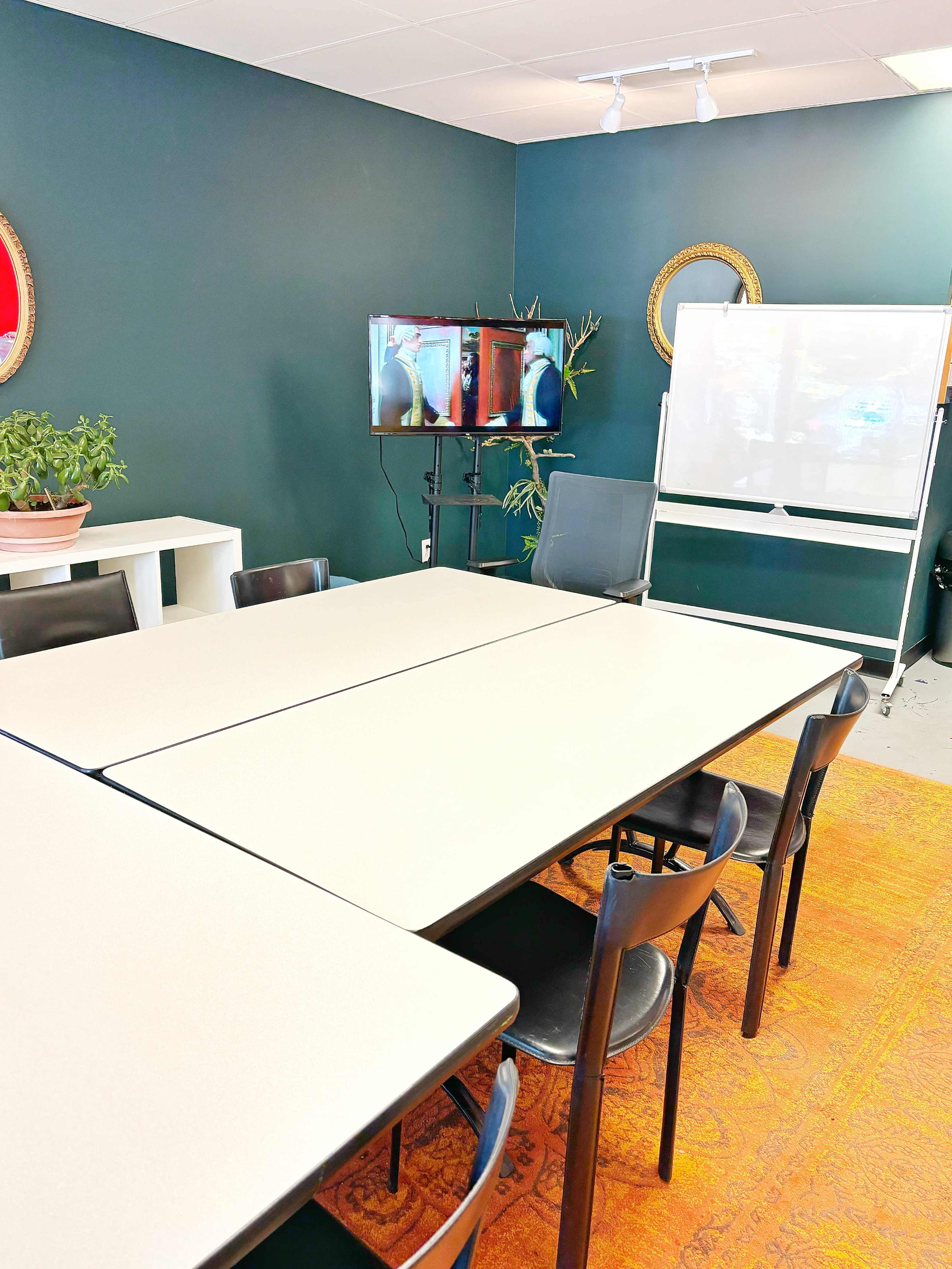 A conference room with a large rectangular table surrounded by black chairs, a whiteboard, and two screens displaying visuals on a green wall.