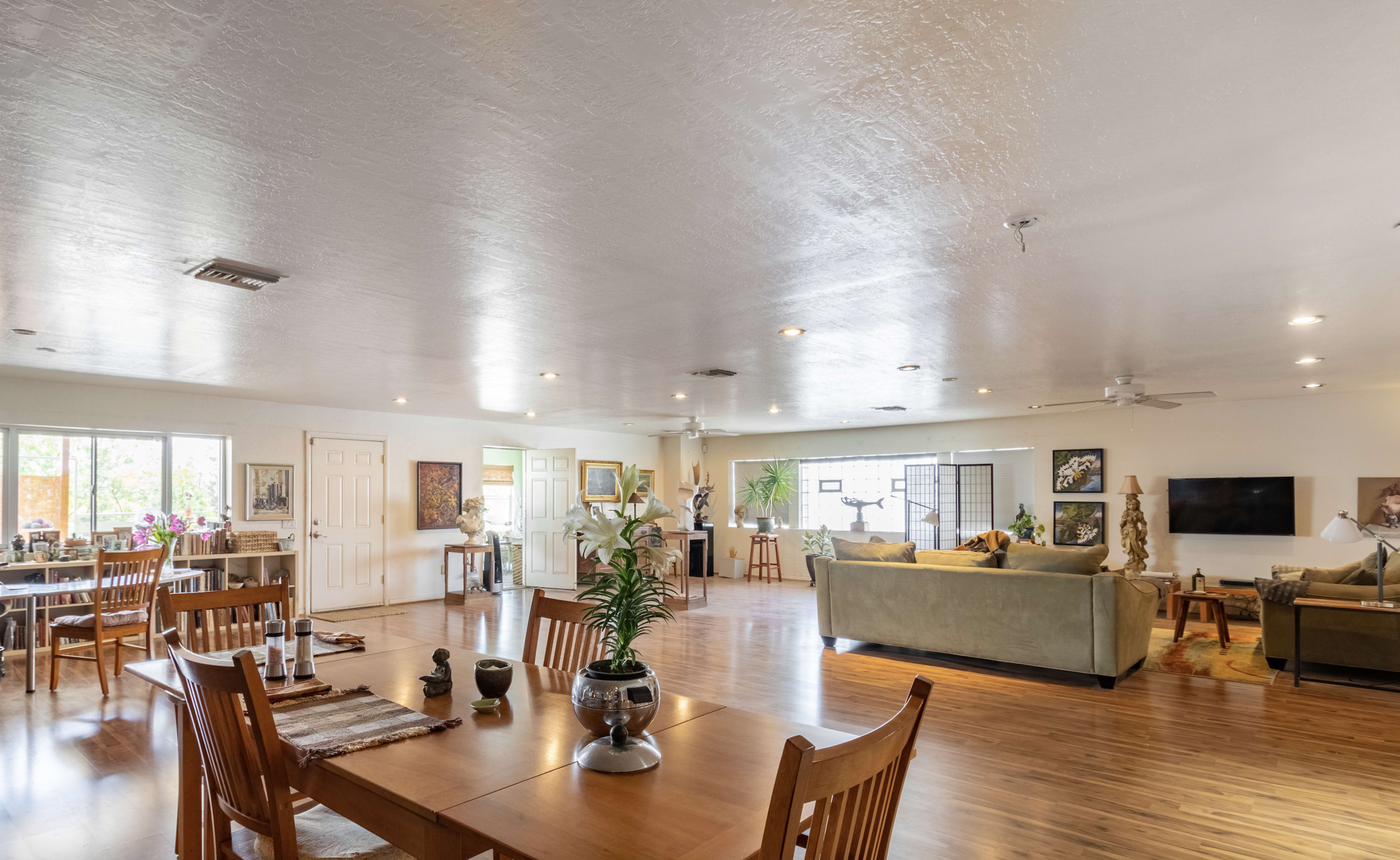 A spacious living area with wooden floors, multiple seating arrangements, and dining tables, illuminated by natural light from large windows.