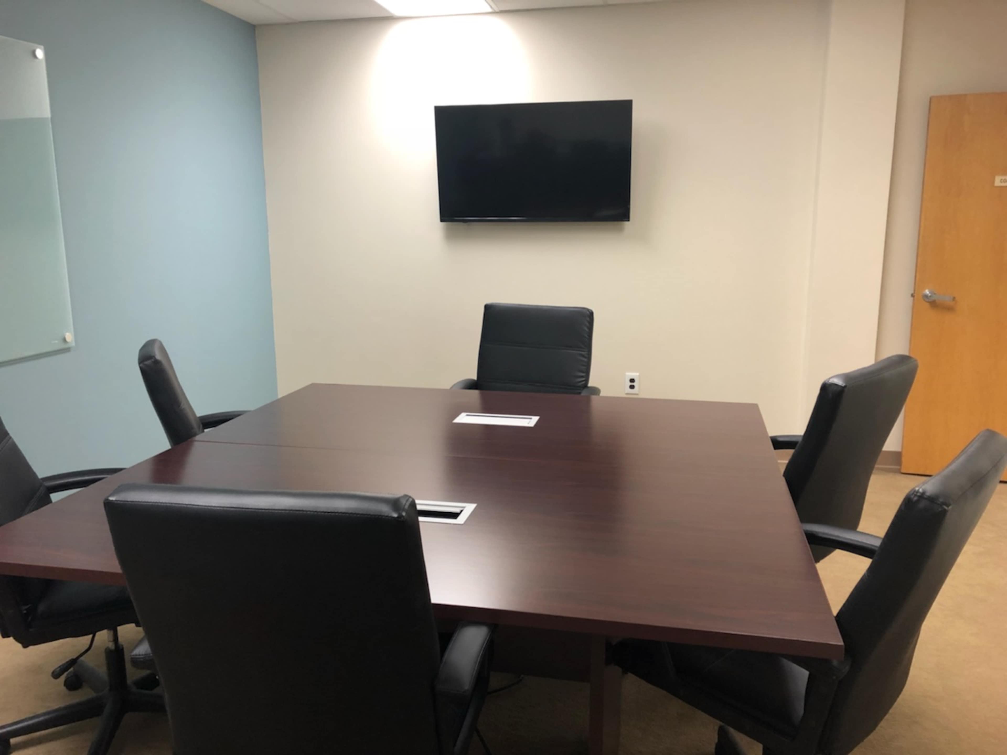 A vacant conference room features a large wooden table surrounded by black leather chairs, with a wall-mounted television and a door in the background.