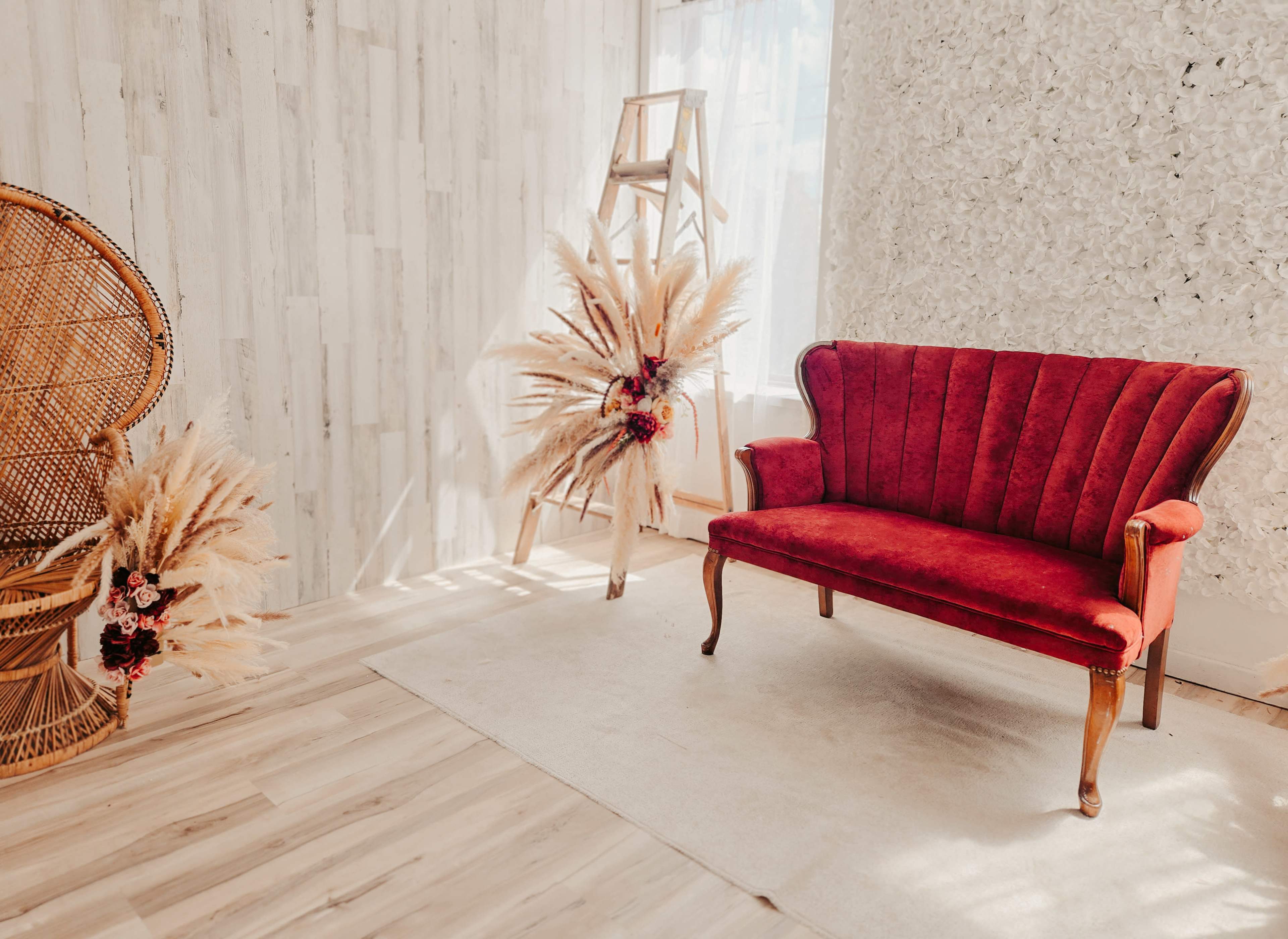 The image shows a cozy interior featuring a red velvet sofa and decorative elements, including dried floral arrangements, against a light-colored wall and wooden flooring.