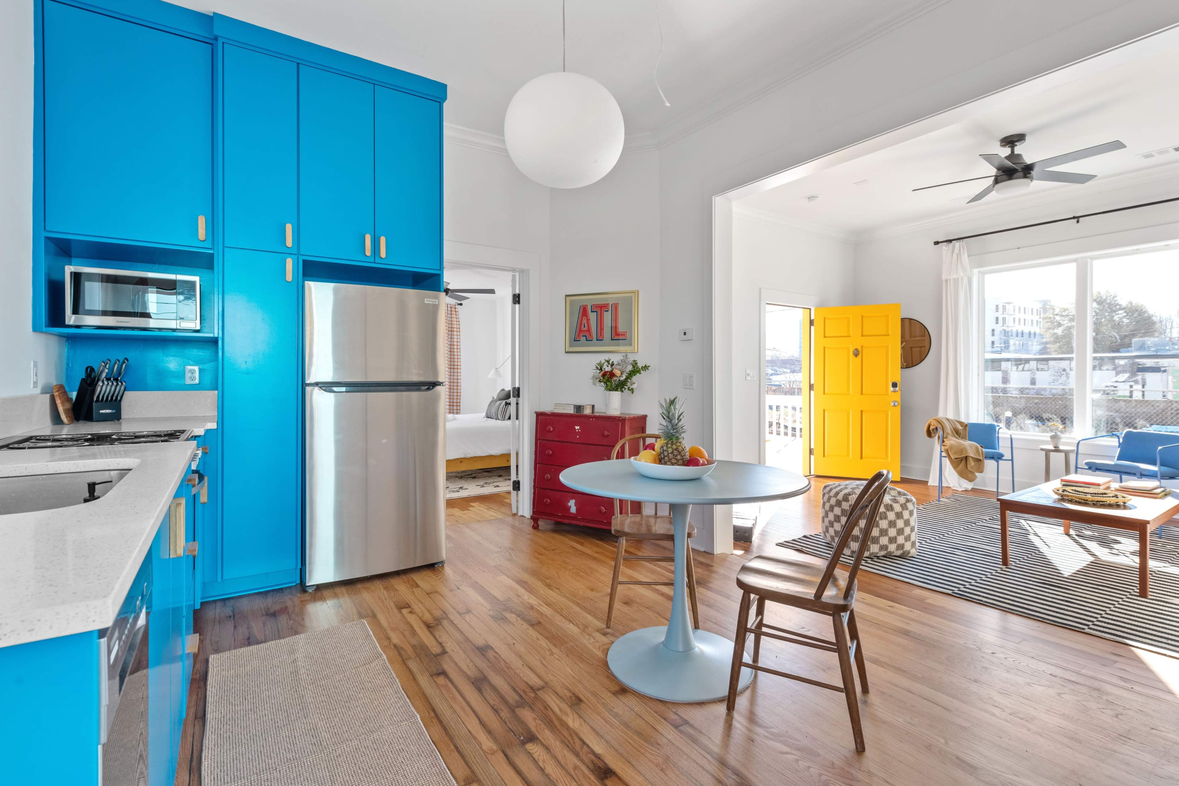 A modern kitchen with bright blue cabinets, stainless steel appliances, and a small dining table next to a large window with a yellow door in the background.