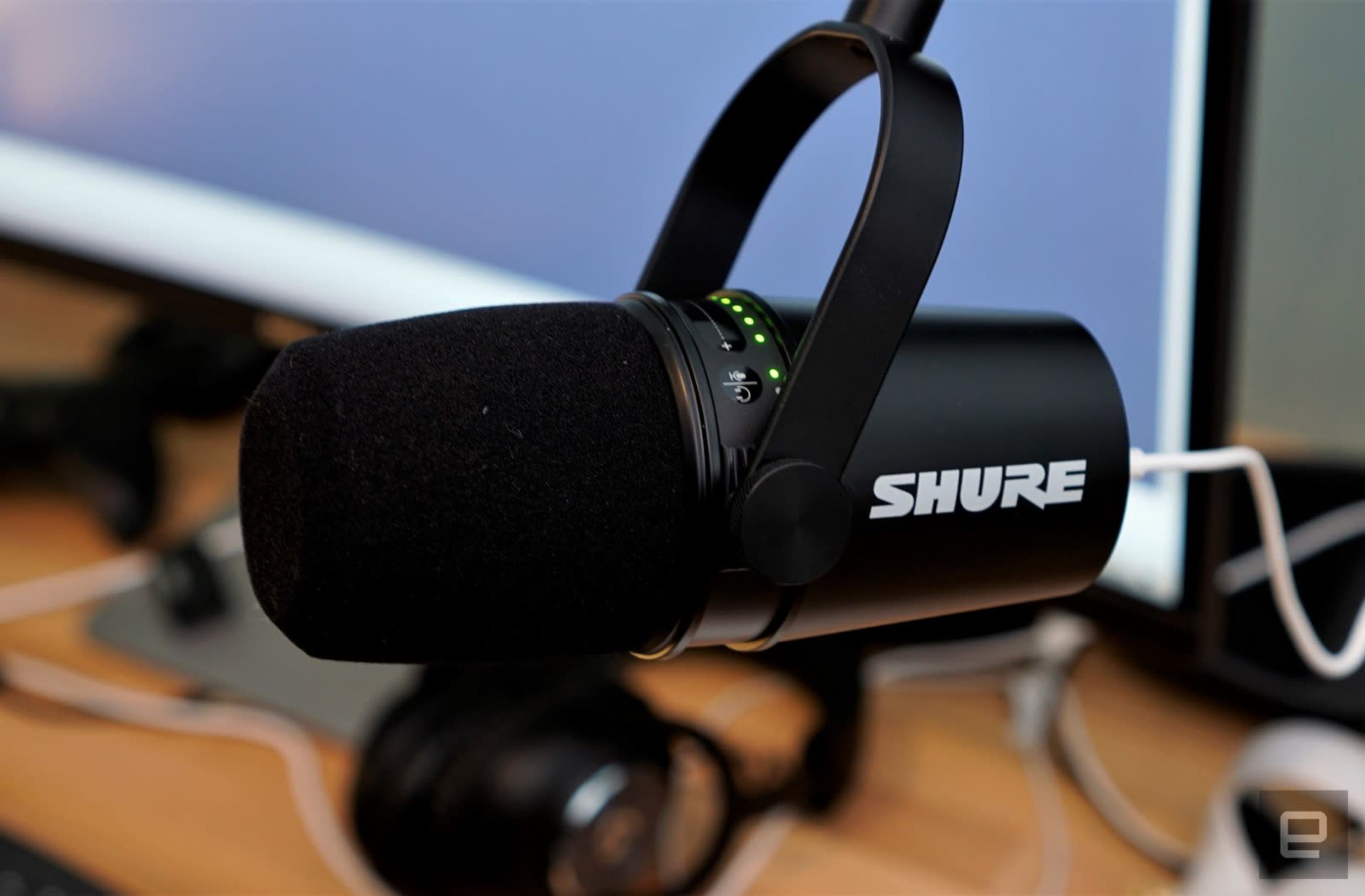 A Shure microphone is mounted on a desk with a computer screen in the background and headphones nearby.