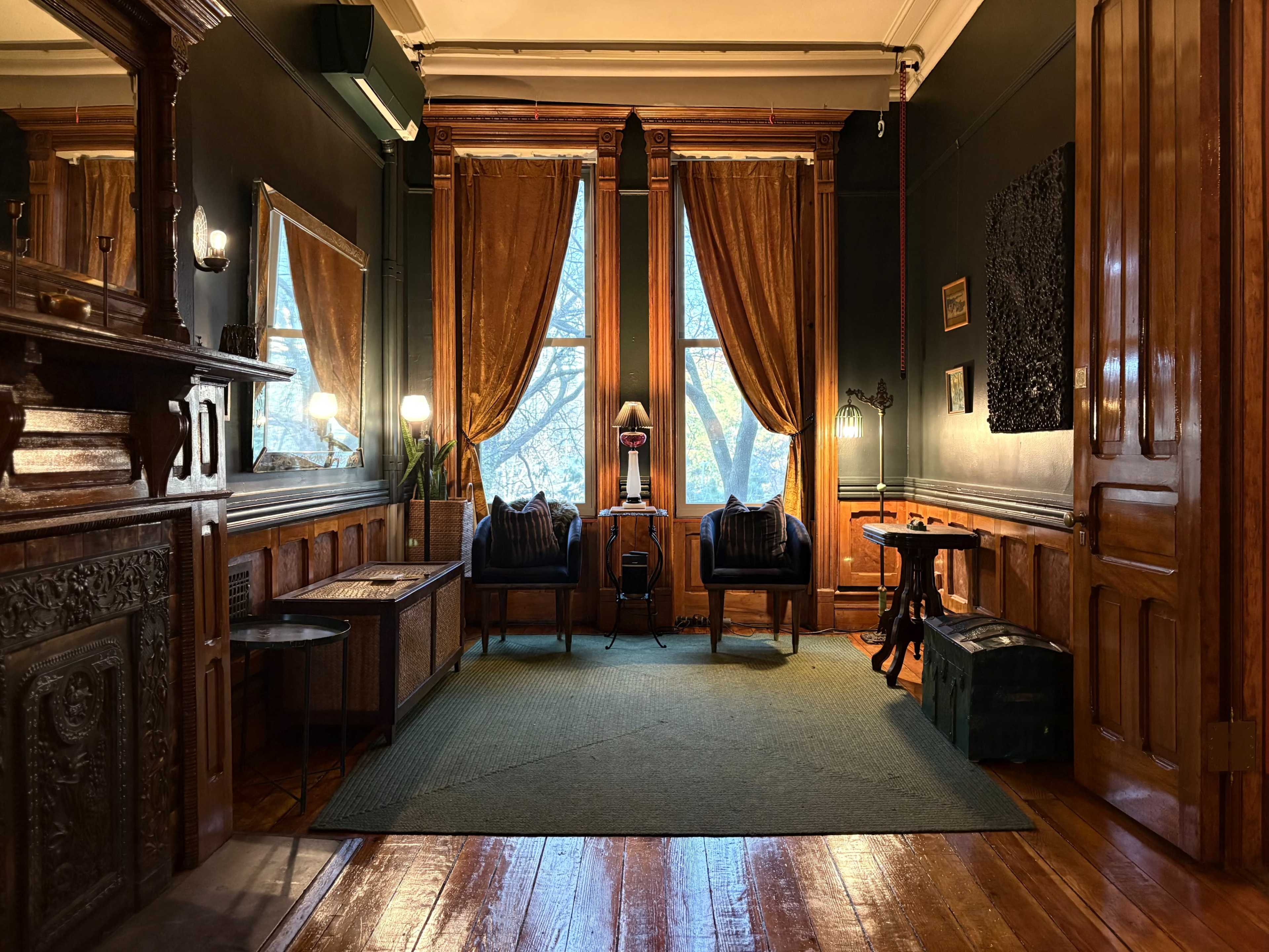 The image shows a cozy room with wooden floors and tall windows draped in orange curtains, featuring two chairs, a small table, and a mirror reflecting the space.