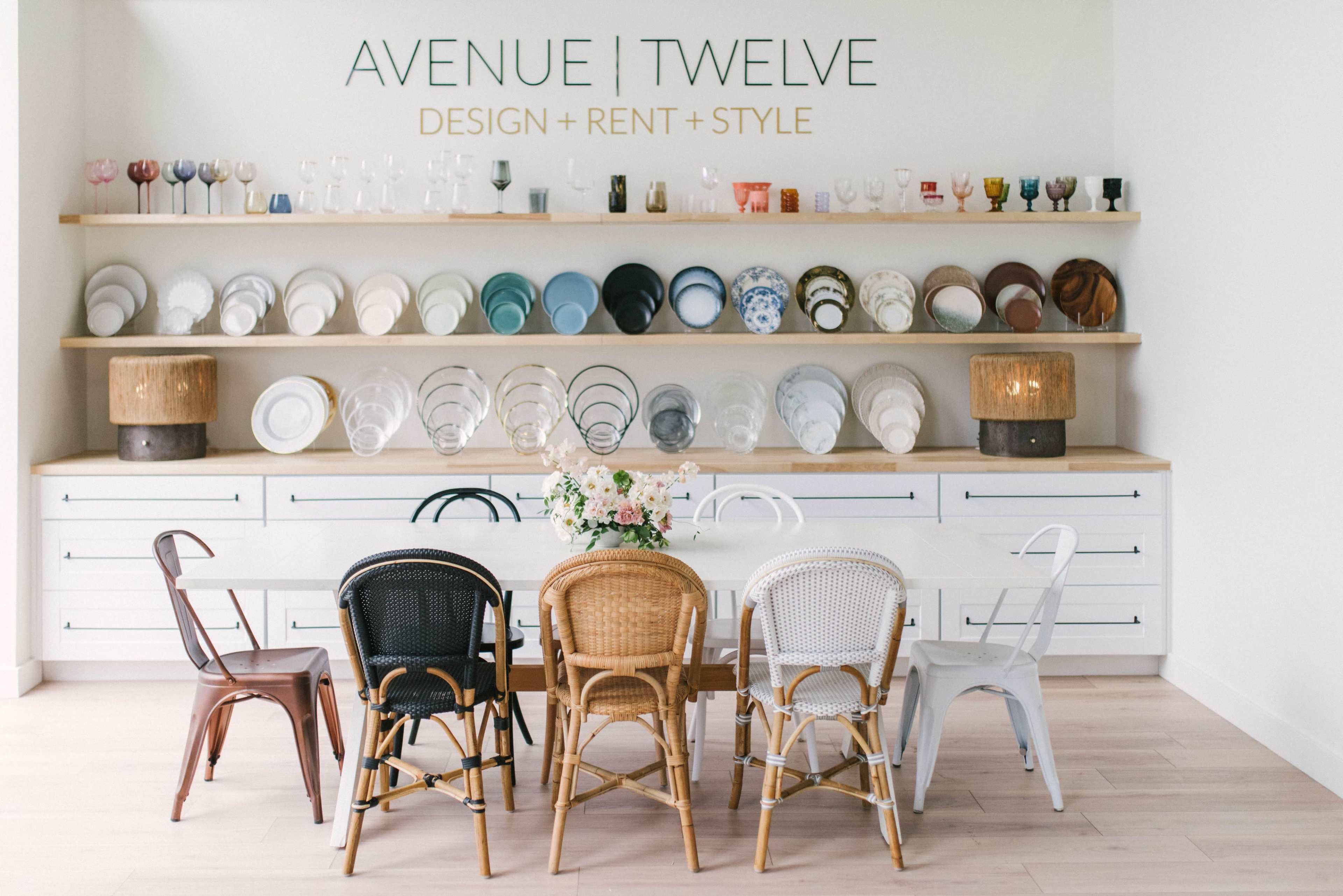 A minimalist interior features a long dining table surrounded by variously colored chairs, with a shelf above displaying an assortment of plates and glassware.