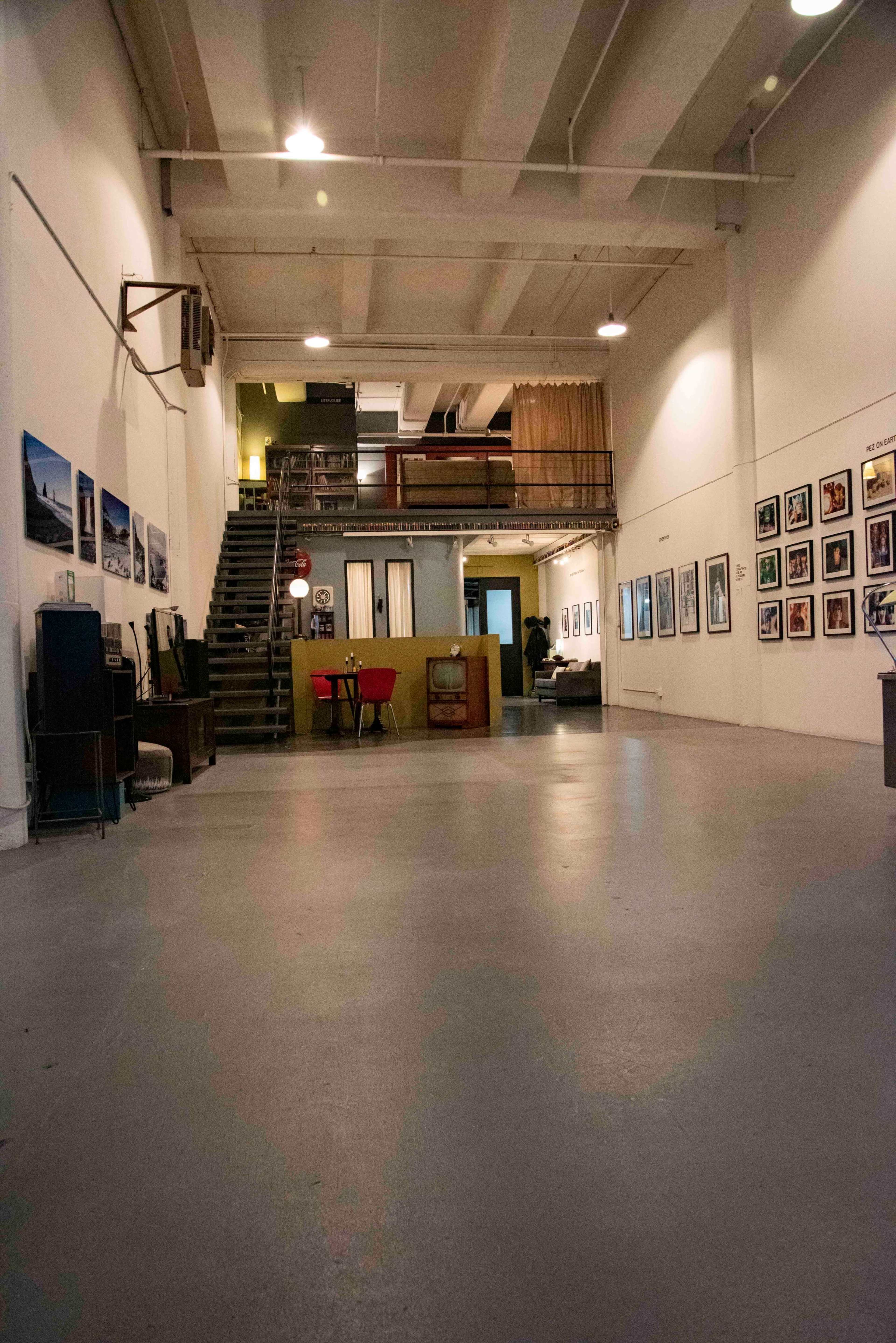 A spacious, minimalist gallery interior with a staircase, framed photographs on the walls, and a few scattered furniture pieces.