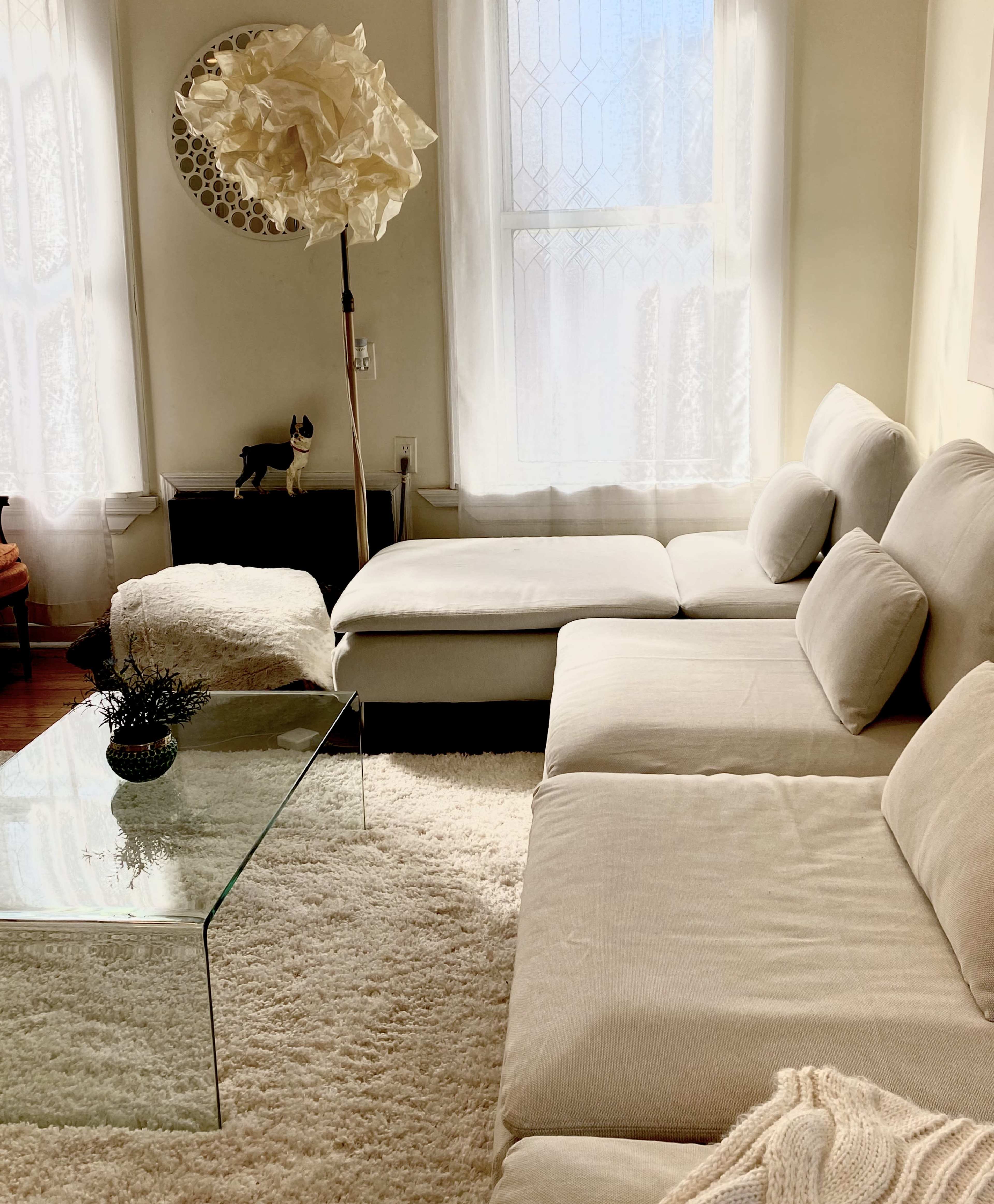 A cozy living room with a light-colored sectional sofa, a glass coffee table, and a textured rug, illuminated by natural light from large windows.
