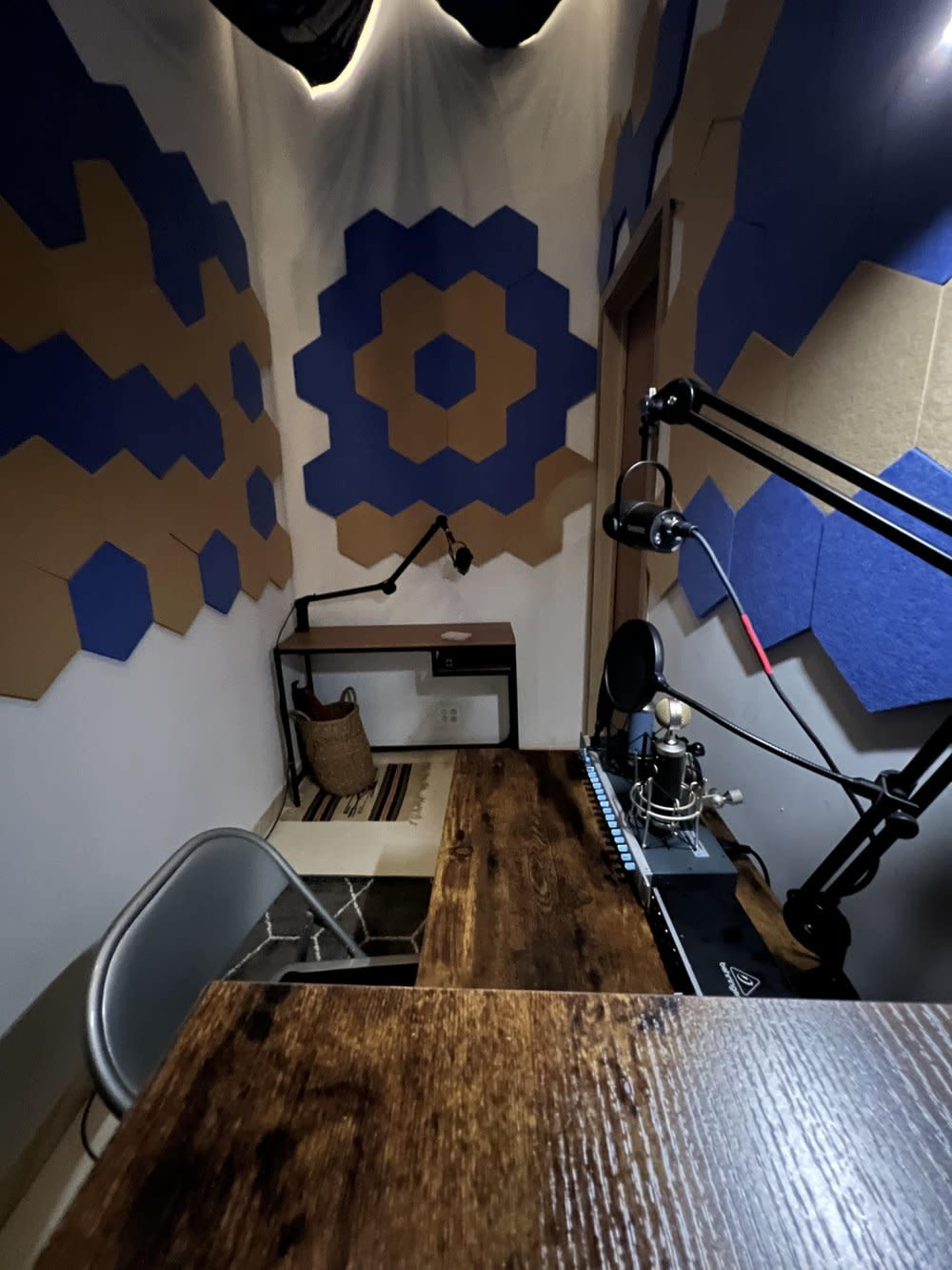 A small recording studio featuring a wooden desk, a microphone setup, and soundproofed walls with blue and tan hexagonal patterns.