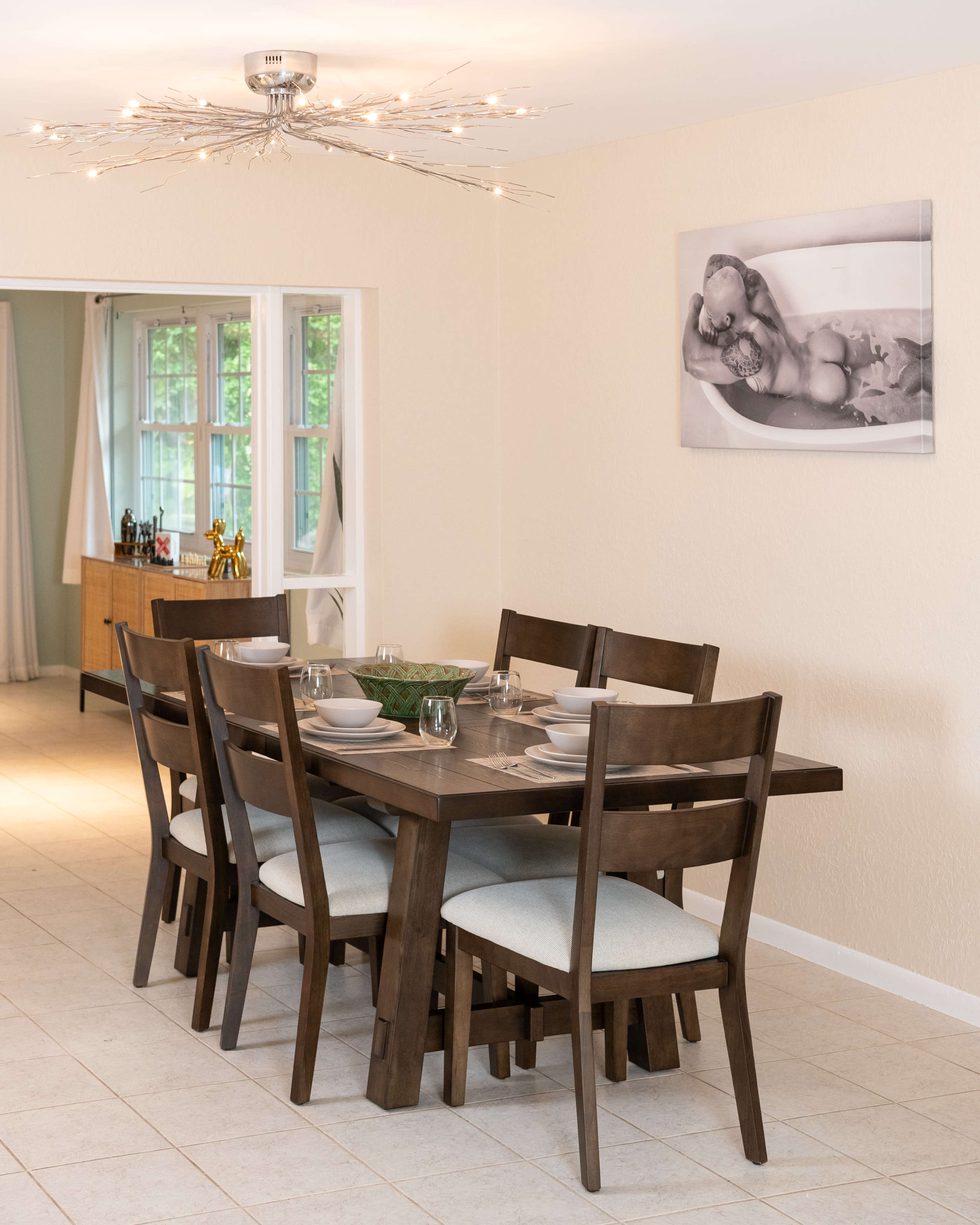 A wooden dining table with six chairs is set for a meal in a brightly lit room with a modern light fixture and a black-and-white photo on the wall.