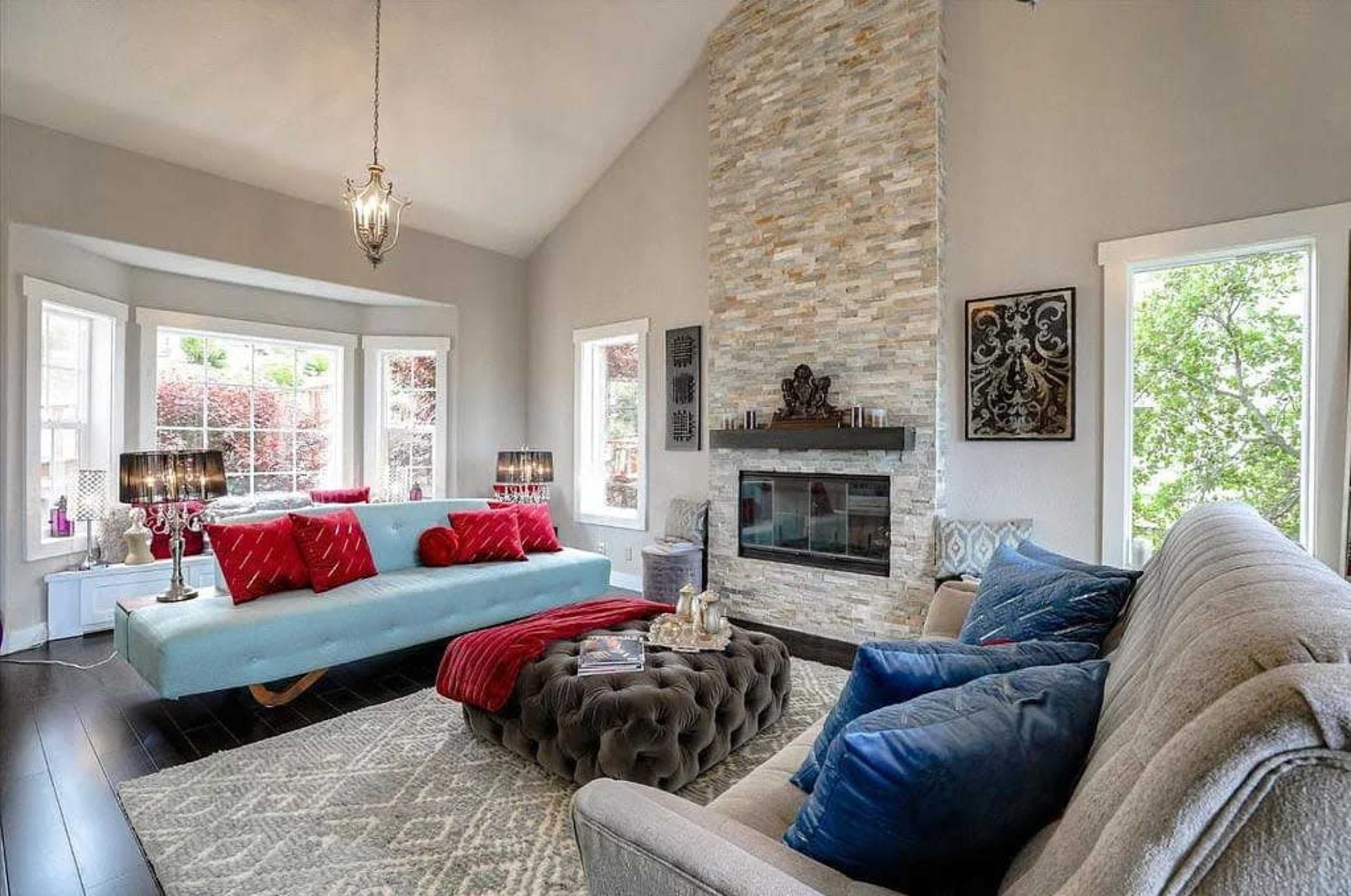 The living room features a light blue sofa and a gray couch arranged around a stone fireplace, with large windows allowing natural light to flood the space.