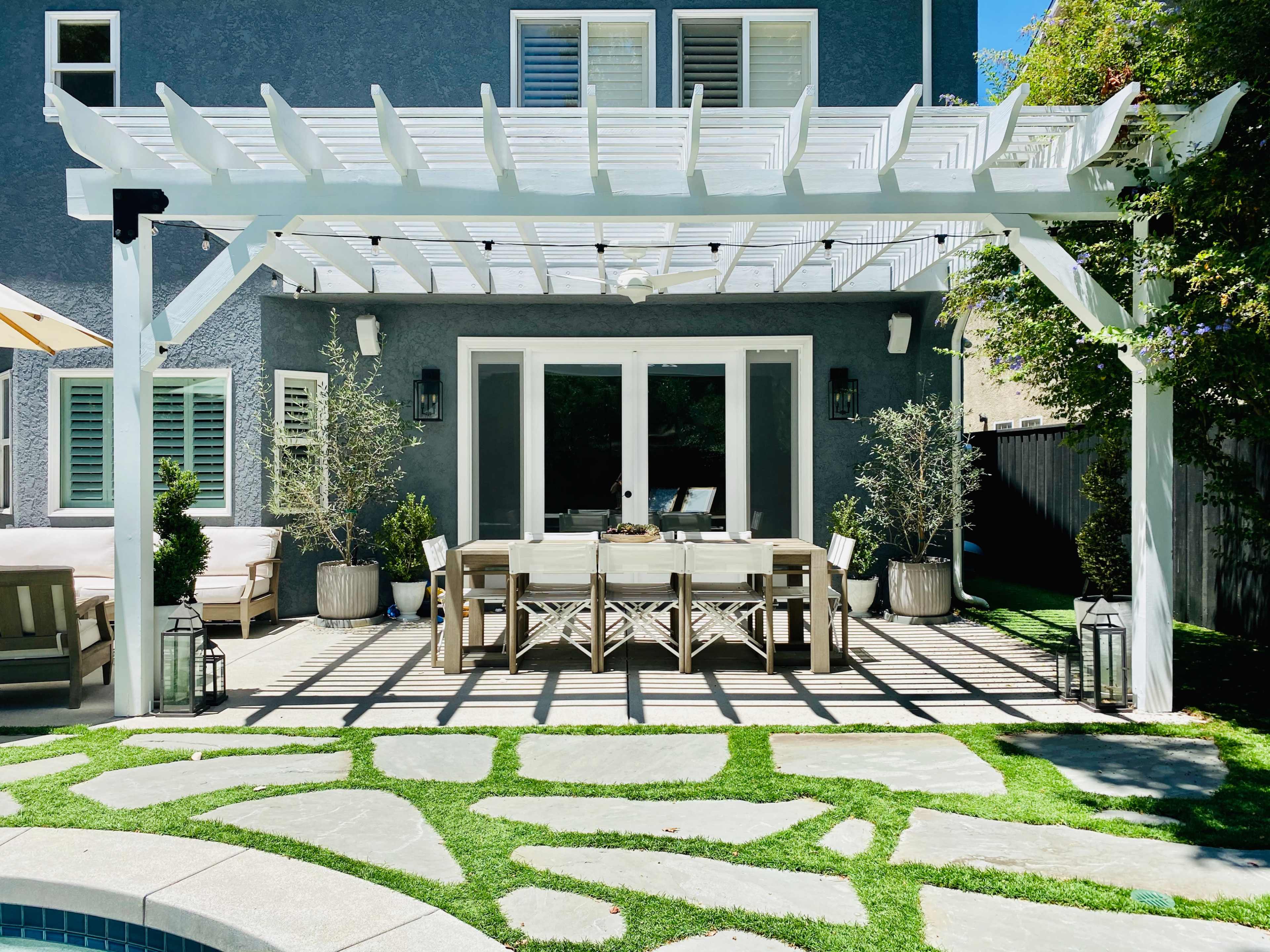 A covered outdoor dining area with a long table and chairs, surrounded by potted plants and decorative lanterns, adjacent to a landscaped stone pathway and green grass.