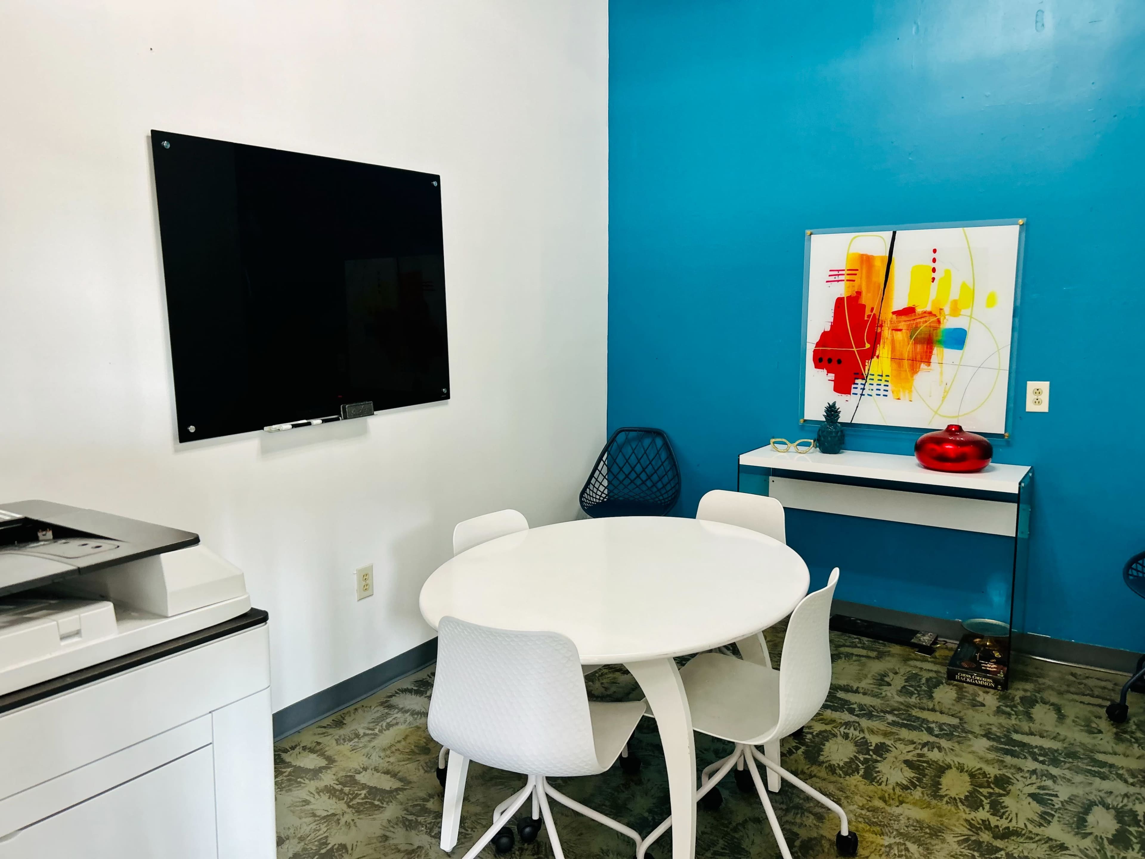A small meeting room features a round white table with four chairs, a black dry-erase board on the wall, and colorful artwork.