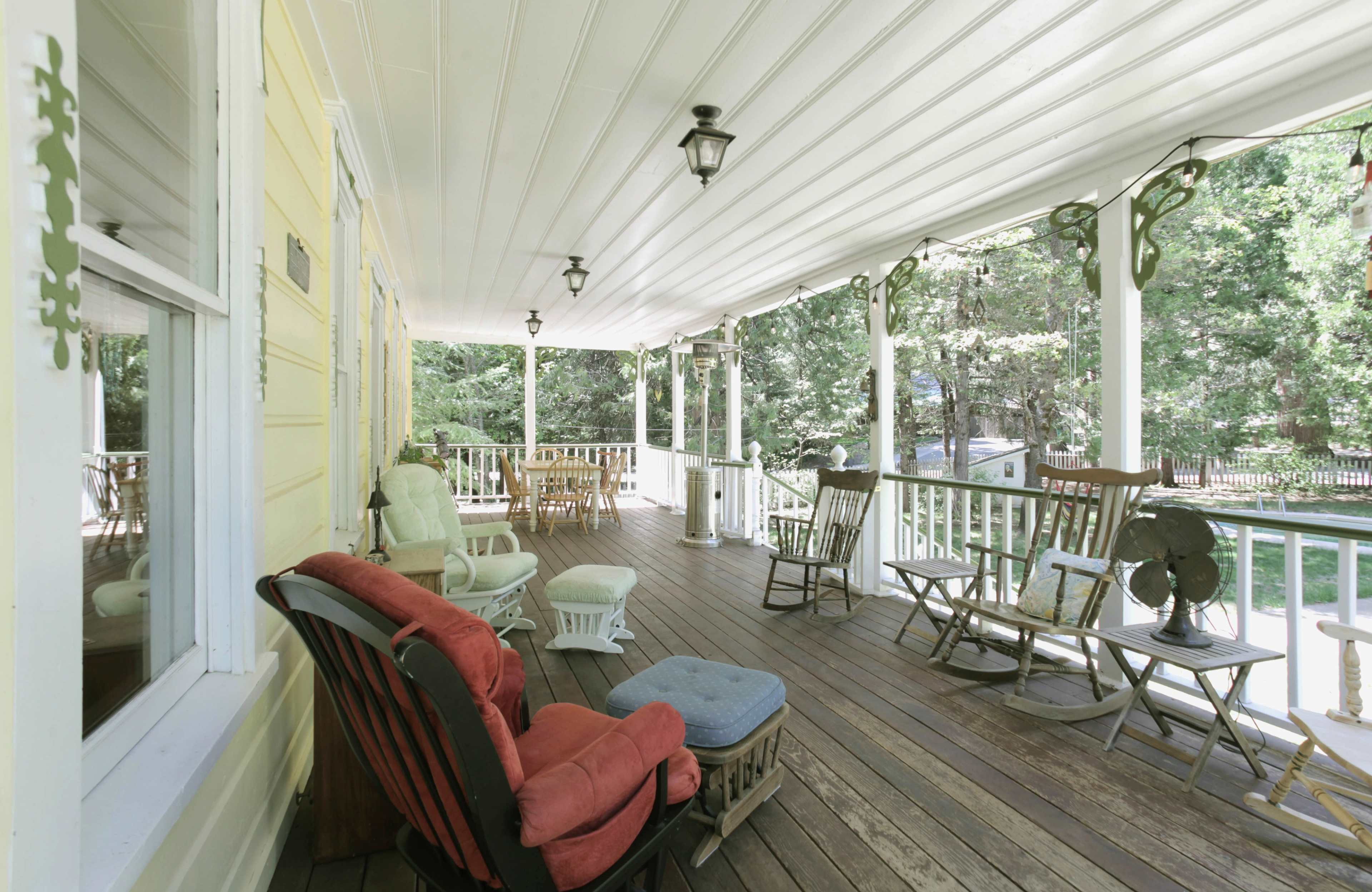 The image shows a spacious porch with various types of seating arranged along a wooden deck, surrounded by trees.