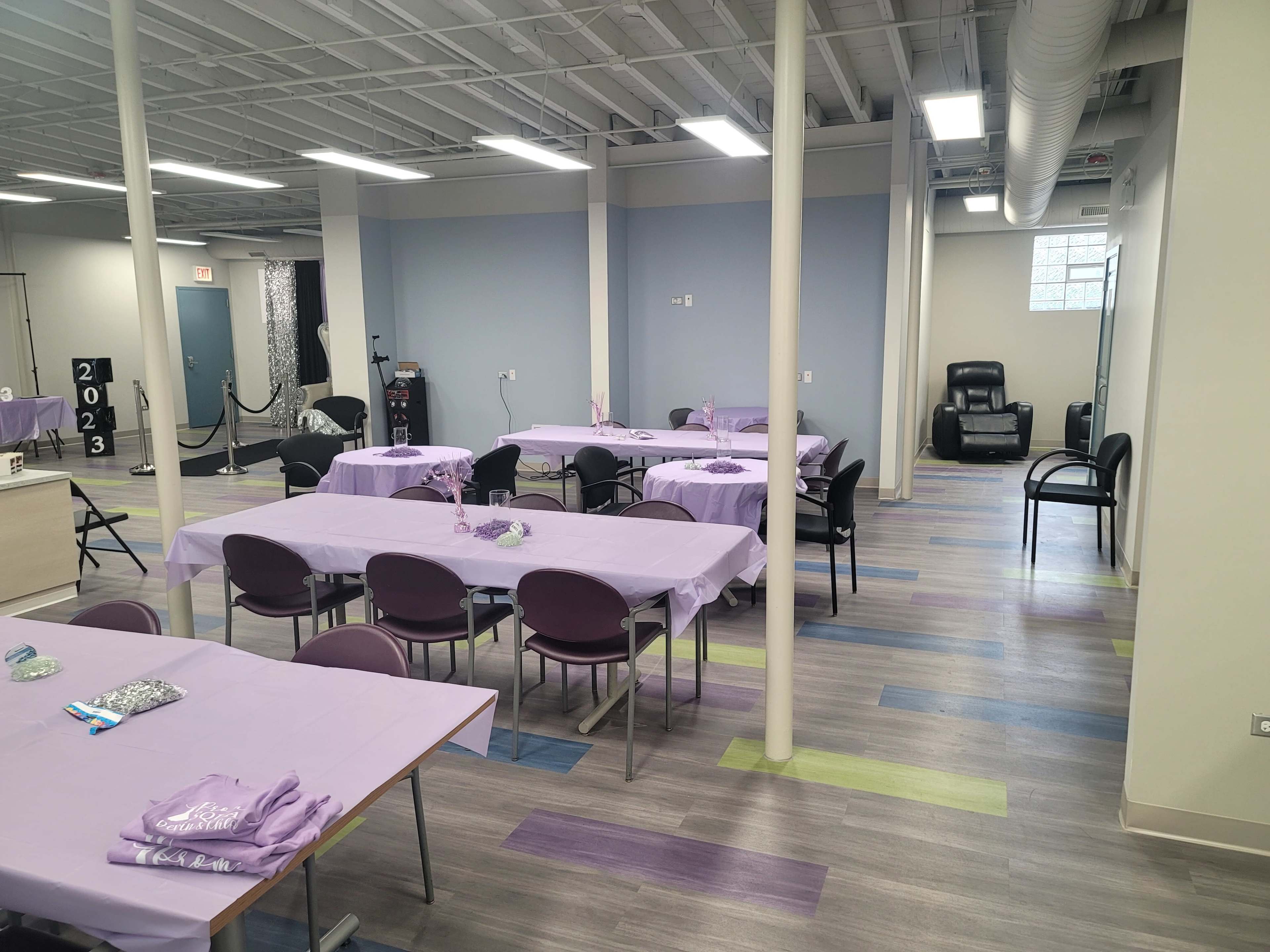 A spacious, well-lit room with several tables covered in purple tablecloths and a few chairs arranged around them, along with a massage chair in the corner.