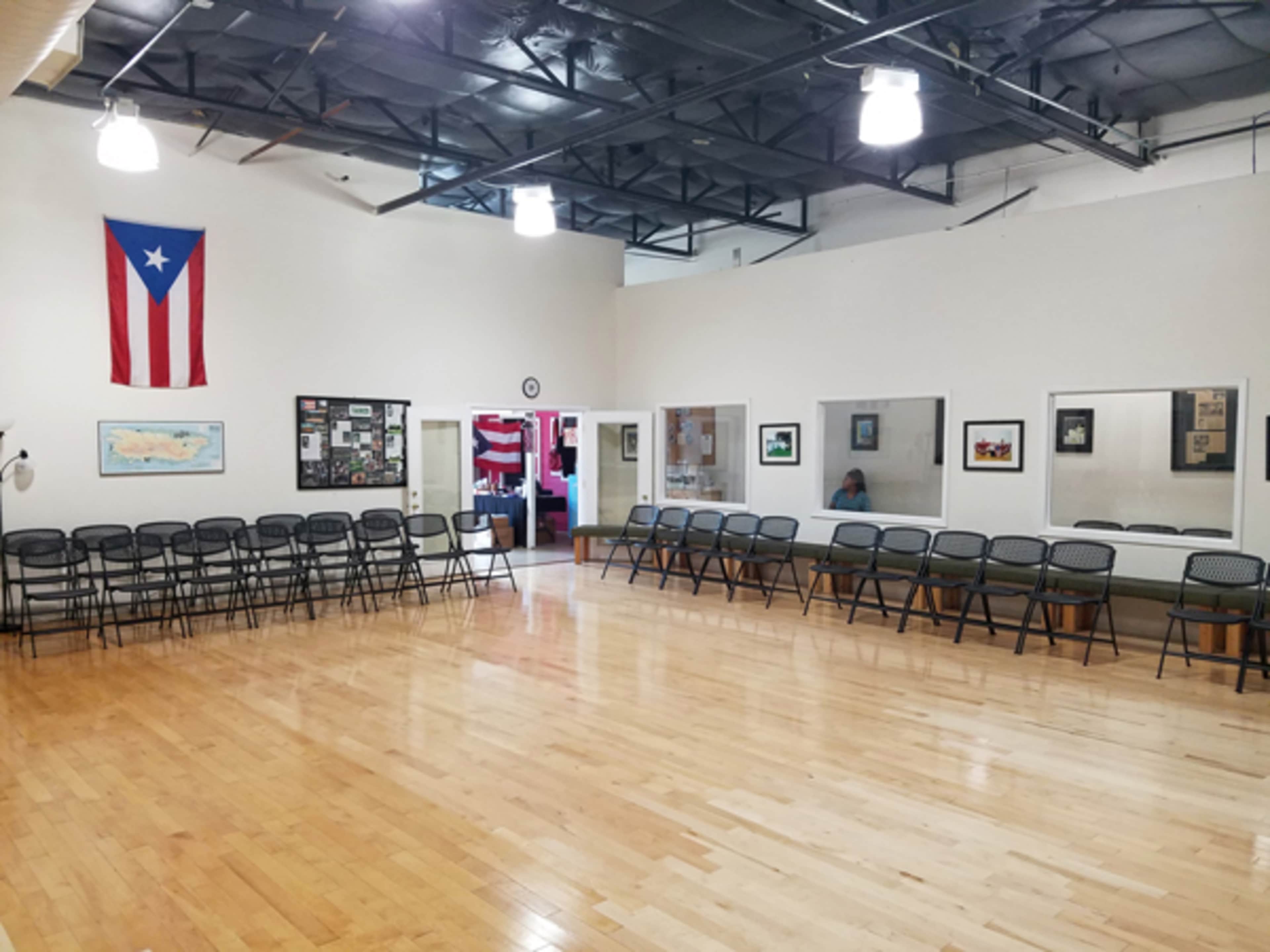 A large, empty room with wooden flooring arranged with rows of metal chairs and featuring a Puerto Rican flag on the wall.