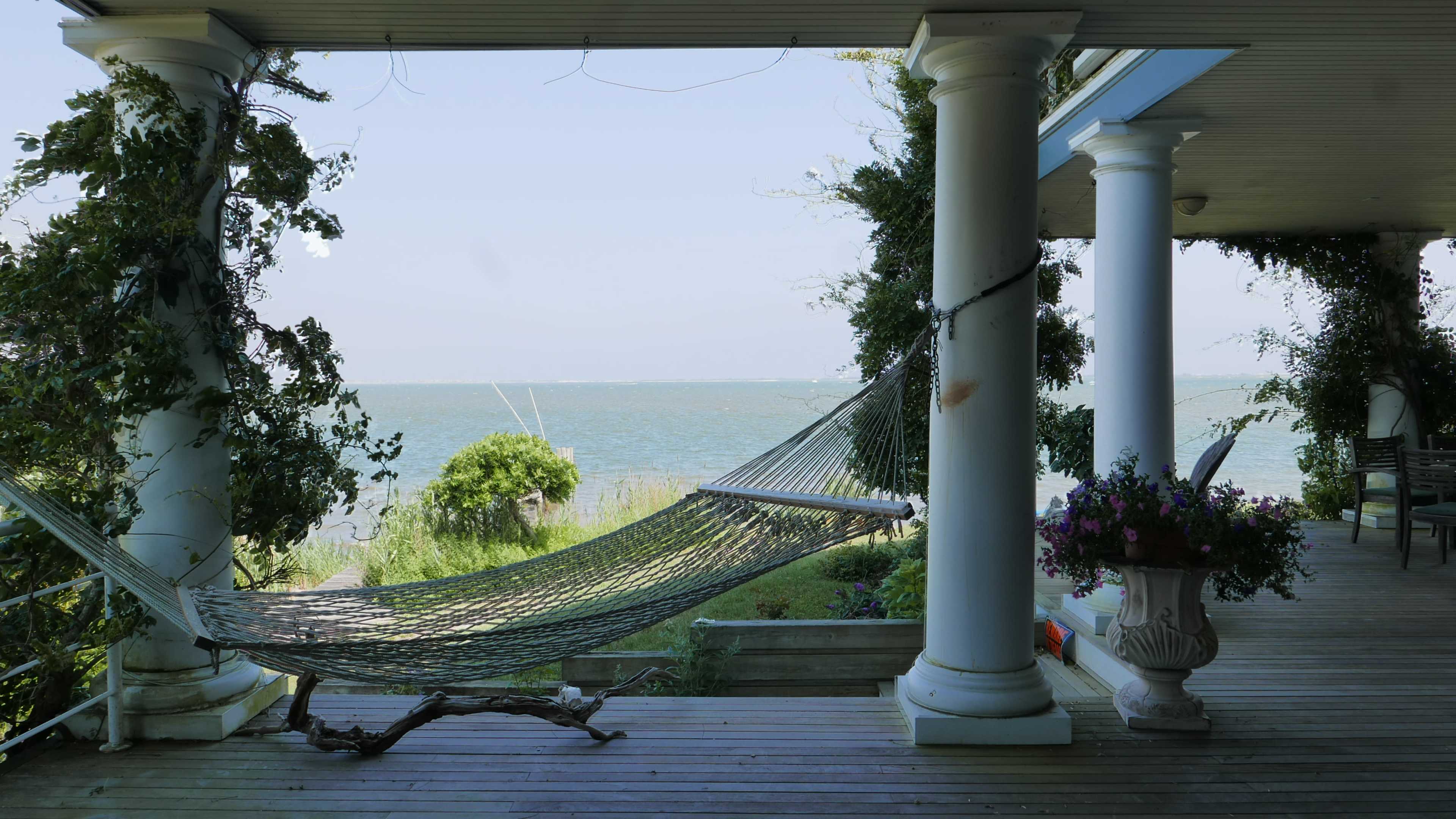 A hammock is suspended between two columns on a porch overlooking a calm body of water.