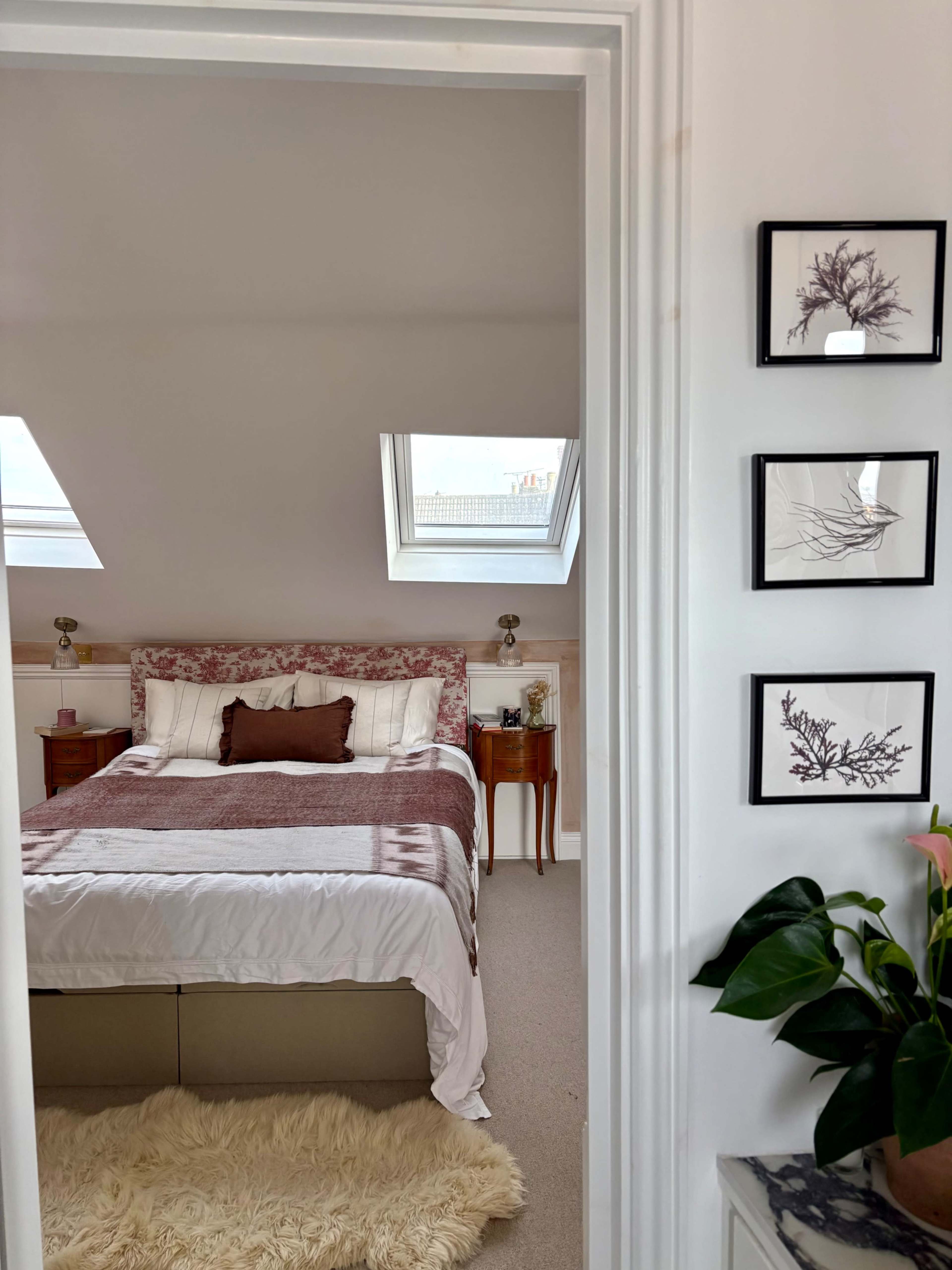 A cozy bedroom features a bed with floral bedding, framed artwork on the wall, and skylights above.