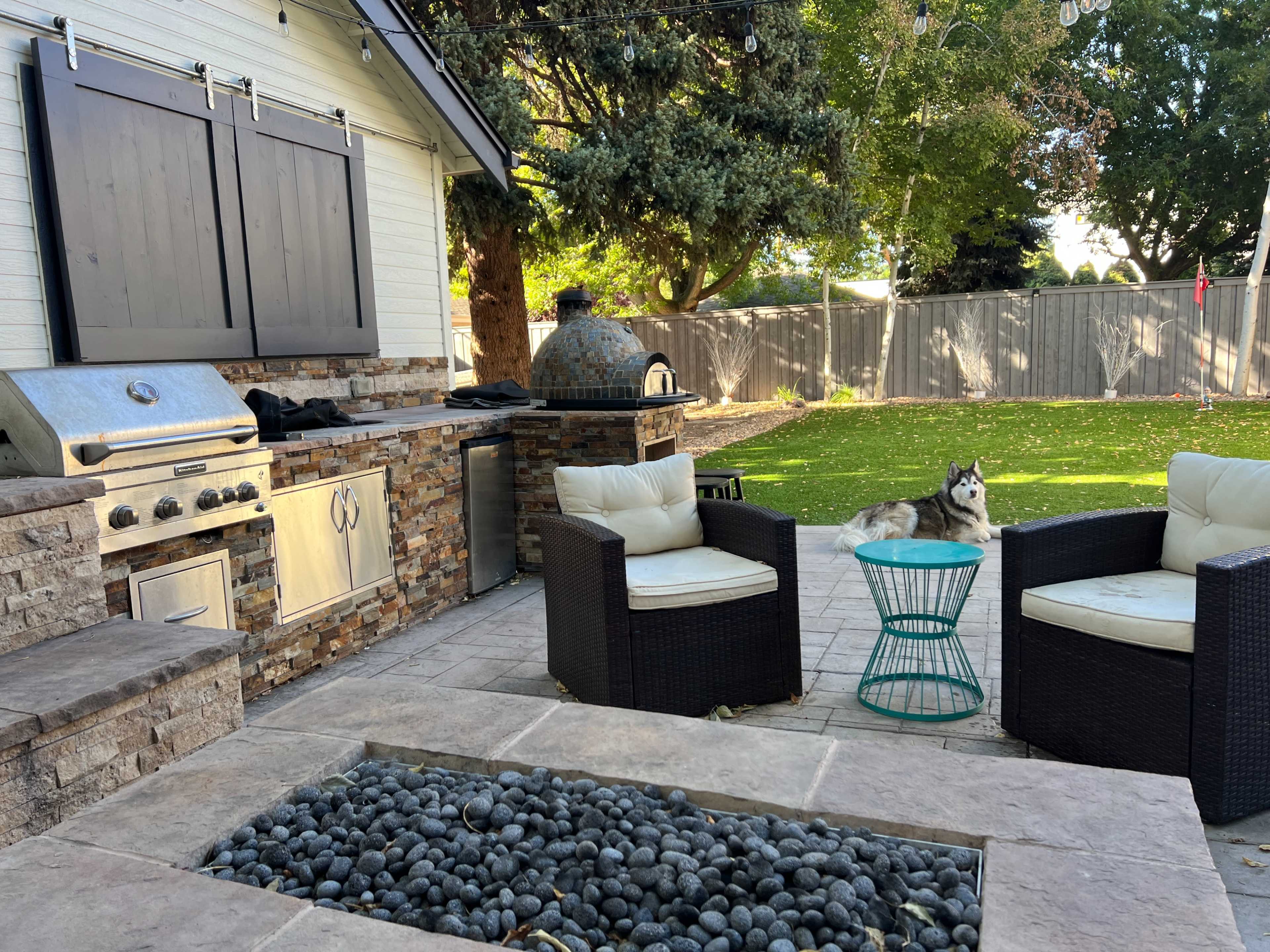The image shows an outdoor kitchen space with a grill, stone seating area, and a dog resting on the grass in a backyard setting.