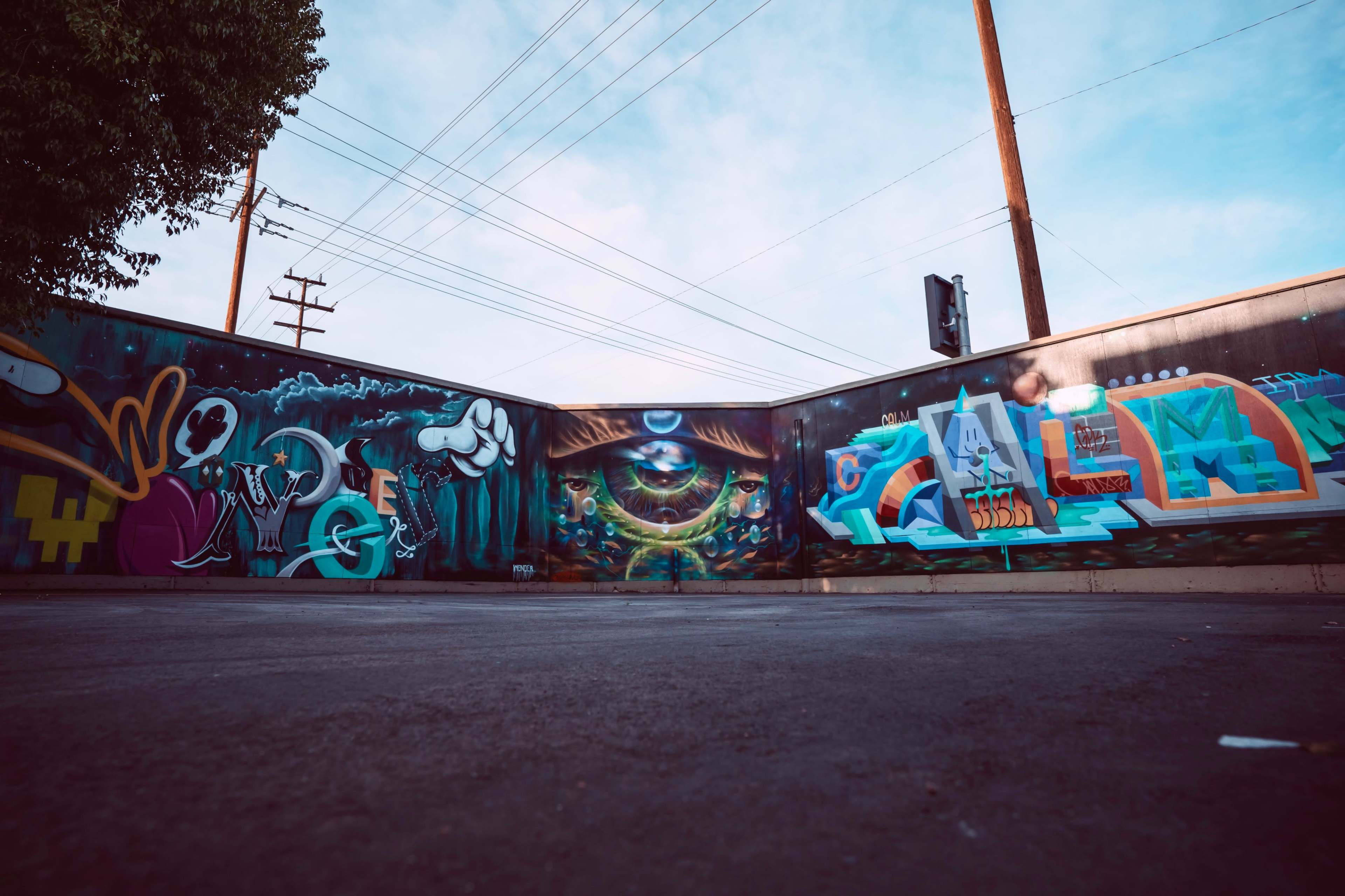 A colorful mural featuring various graffiti styles on a concrete wall at an urban location, with power lines and a traffic light visible in the background.
