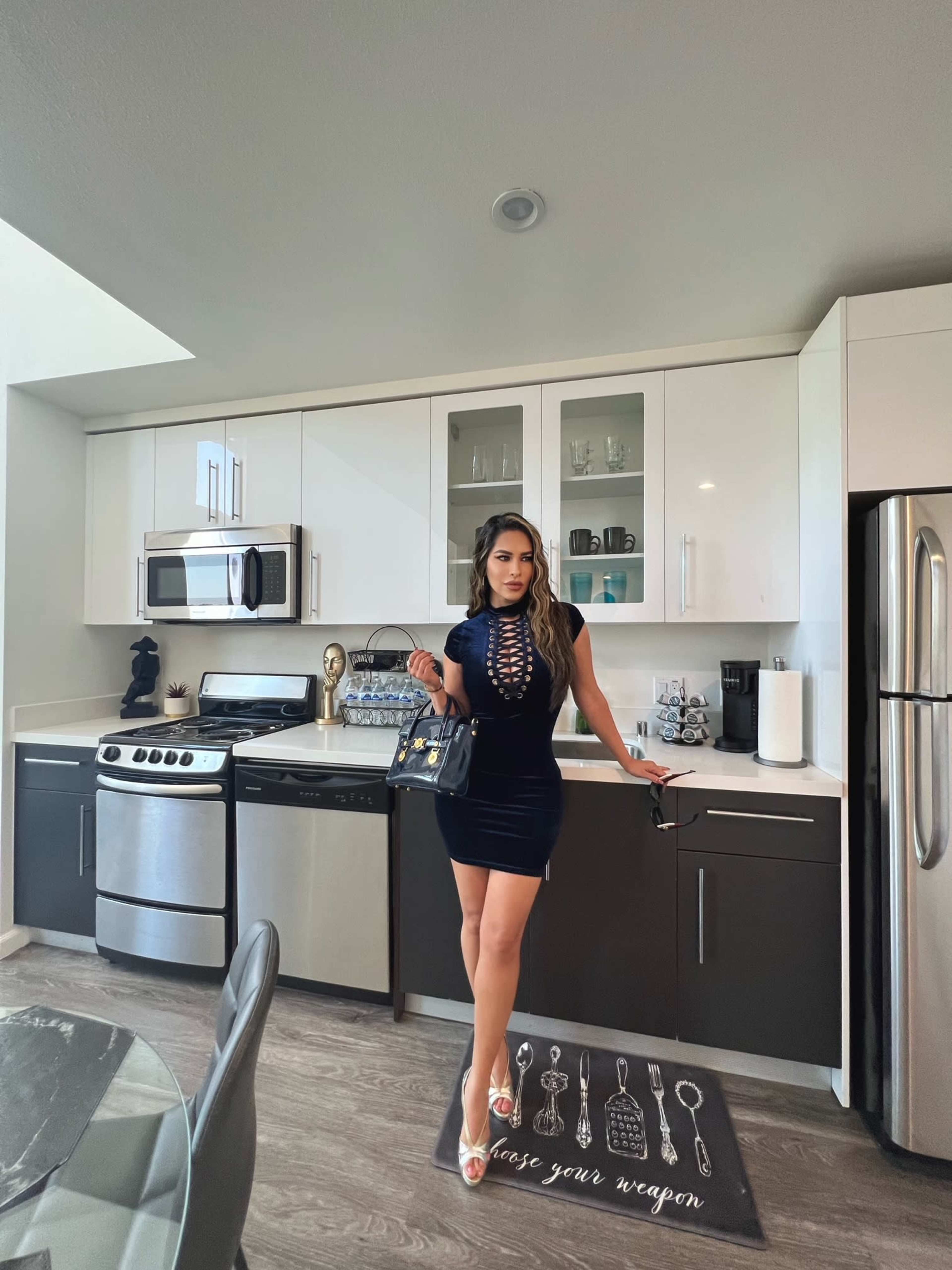 A woman in a black dress stands in a modern kitchen with stainless steel appliances and white cabinets, holding a handbag.