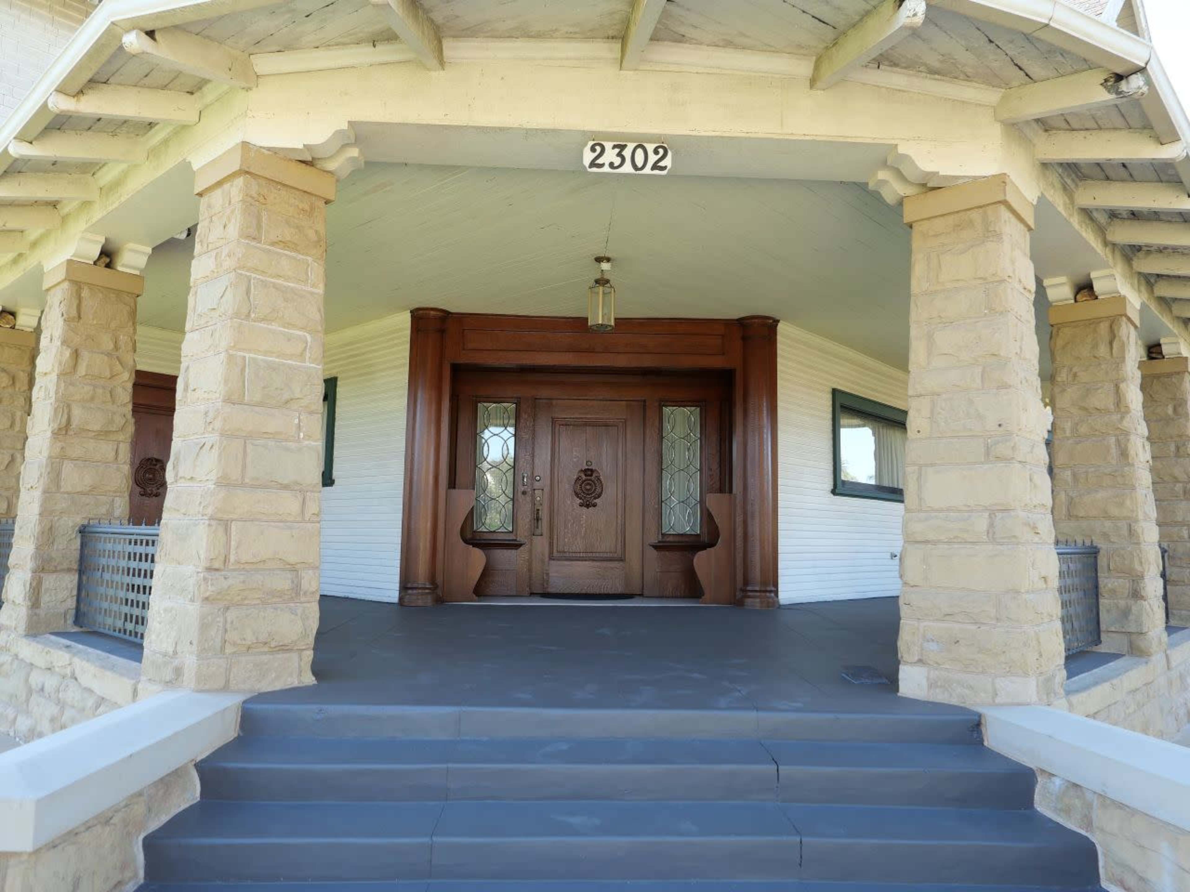 The image shows the entrance of a large house with a stone porch, featuring prominent wooden double doors and decorative glass panels.