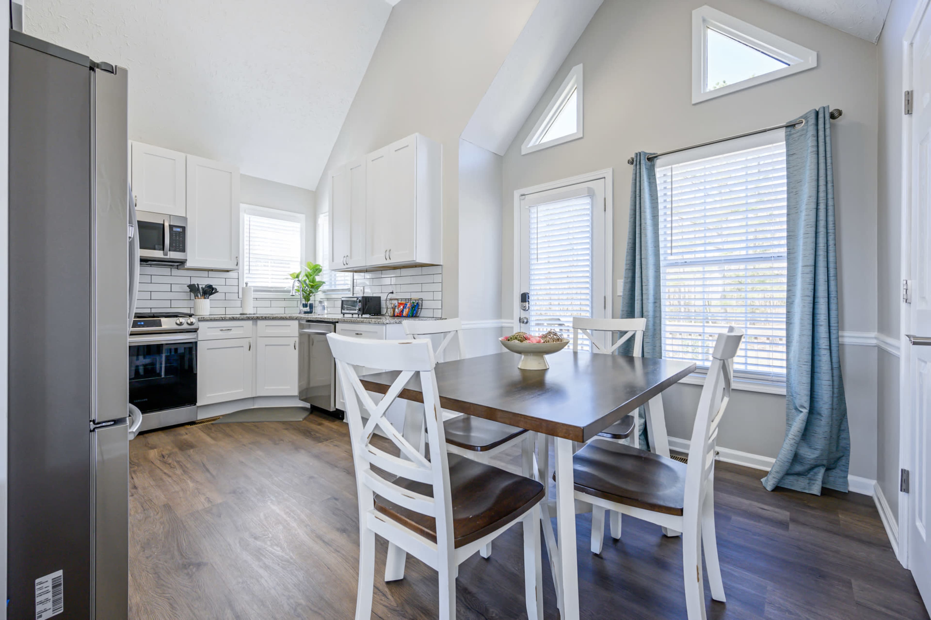 A bright kitchen features white cabinetry, a wooden dining table with four chairs, and large windows allowing natural light to fill the space.