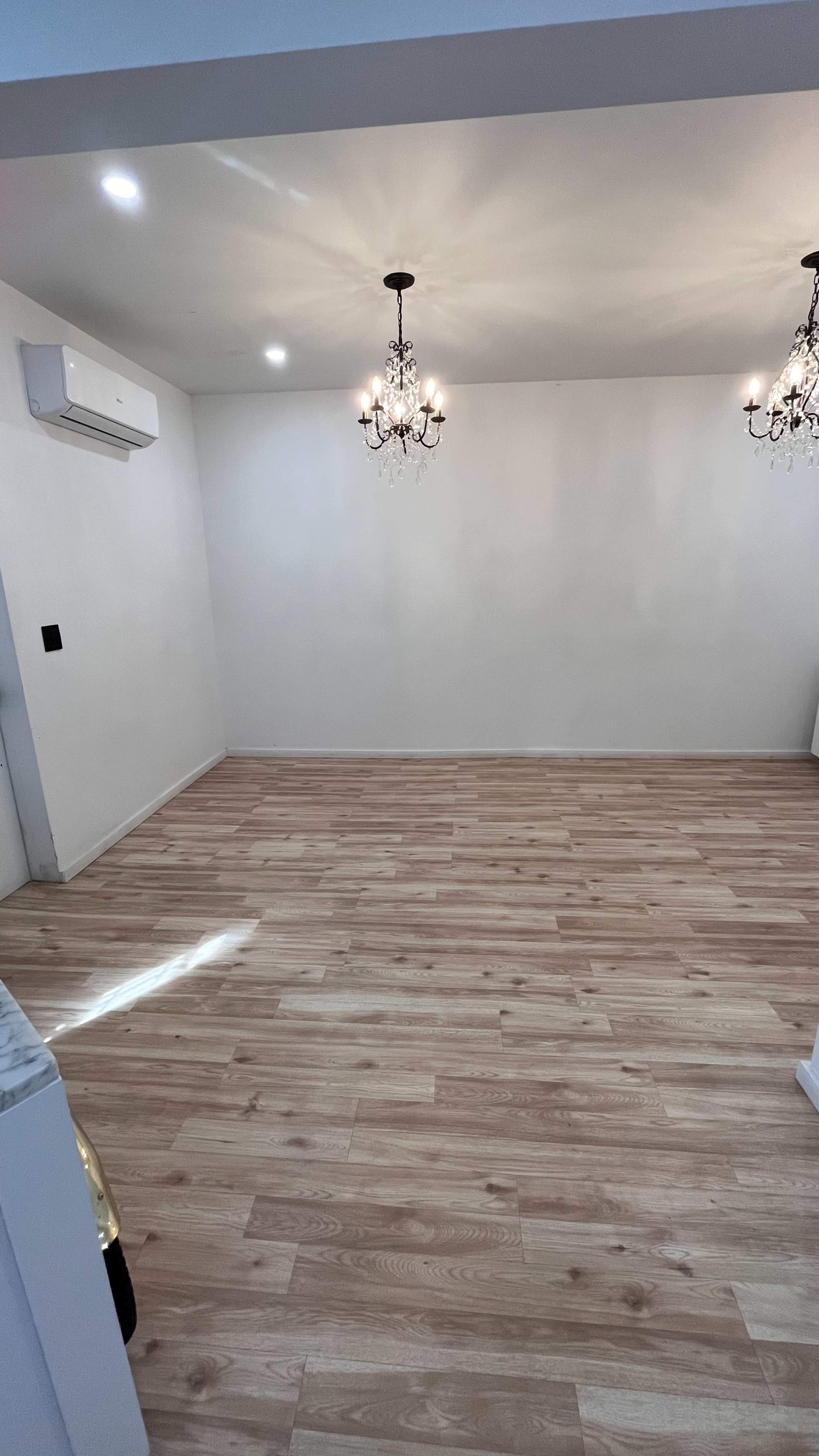 An empty room with light wood flooring, white walls, and two chandeliers hanging from the ceiling.