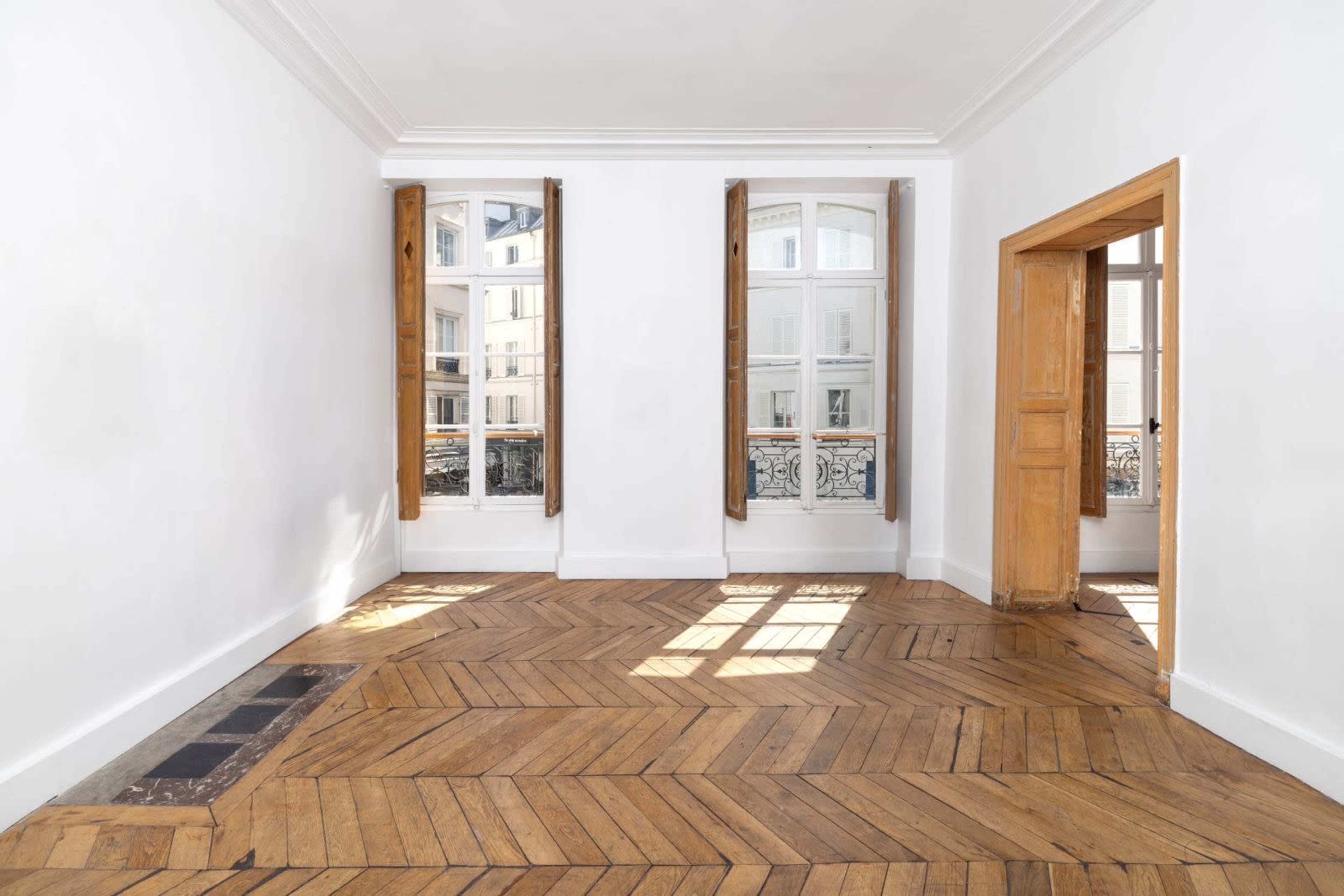 A brightly lit room features wooden flooring, two windows with open shutters, and a door on one side.