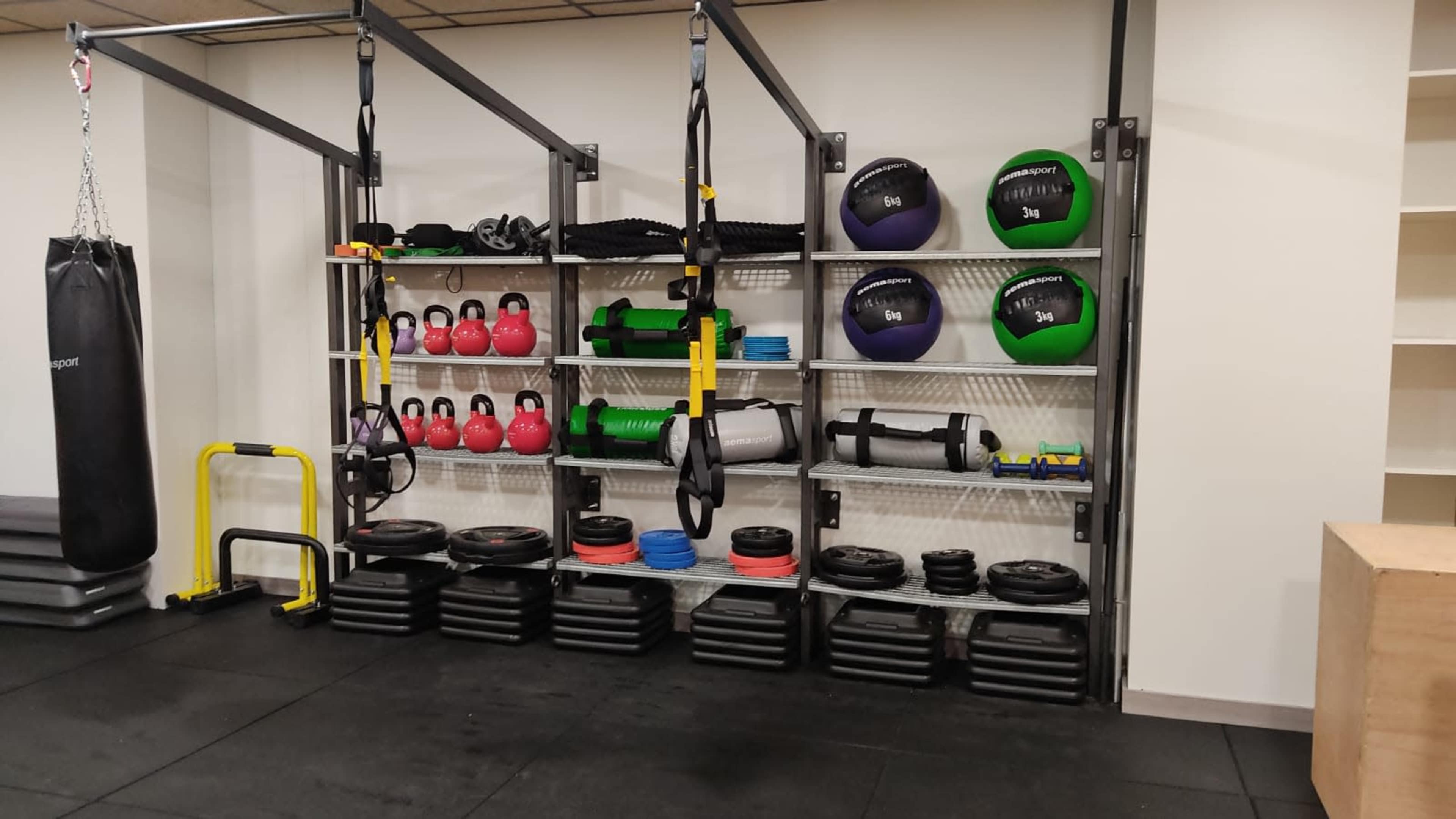 The image shows a gym area featuring various fitness equipment, including kettlebells, medicine balls, resistance bands, and weight plates organized on racks against a wall.