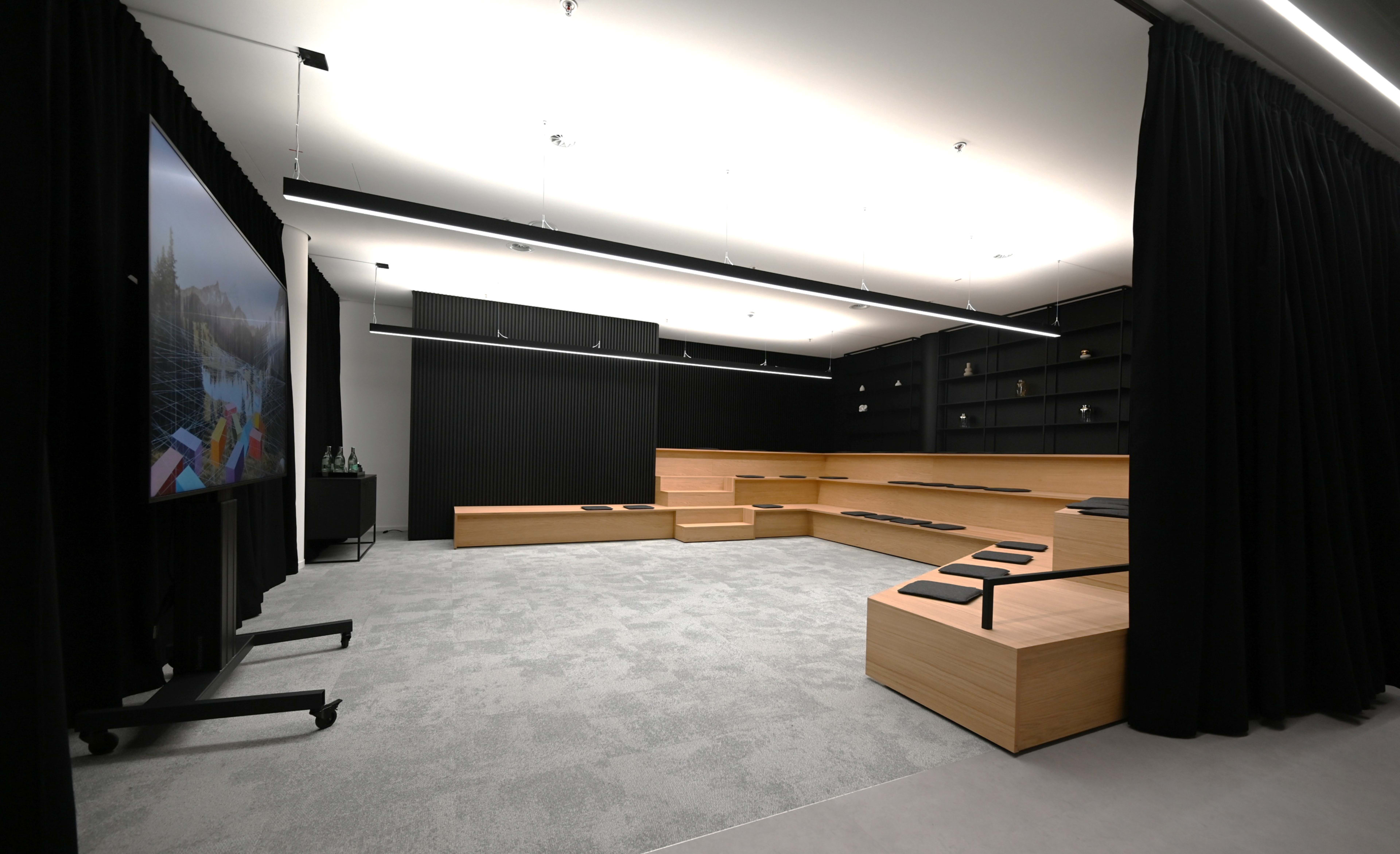 A modern, minimalist meeting room with wooden seating, a large screen, and dark curtains.