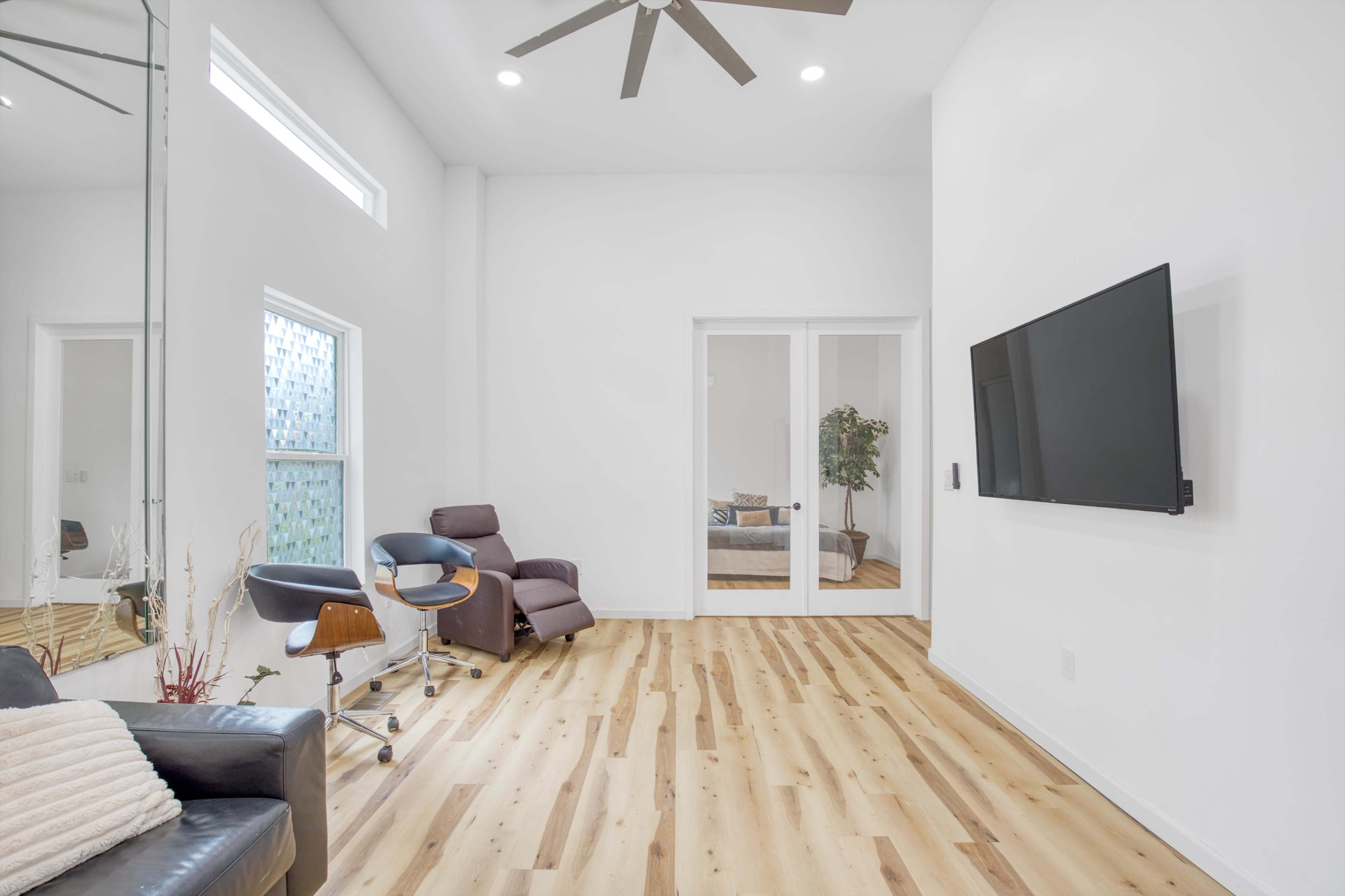 A modern living room features a comfortable chair, a wall-mounted television, and large windows with natural light illuminating the space.