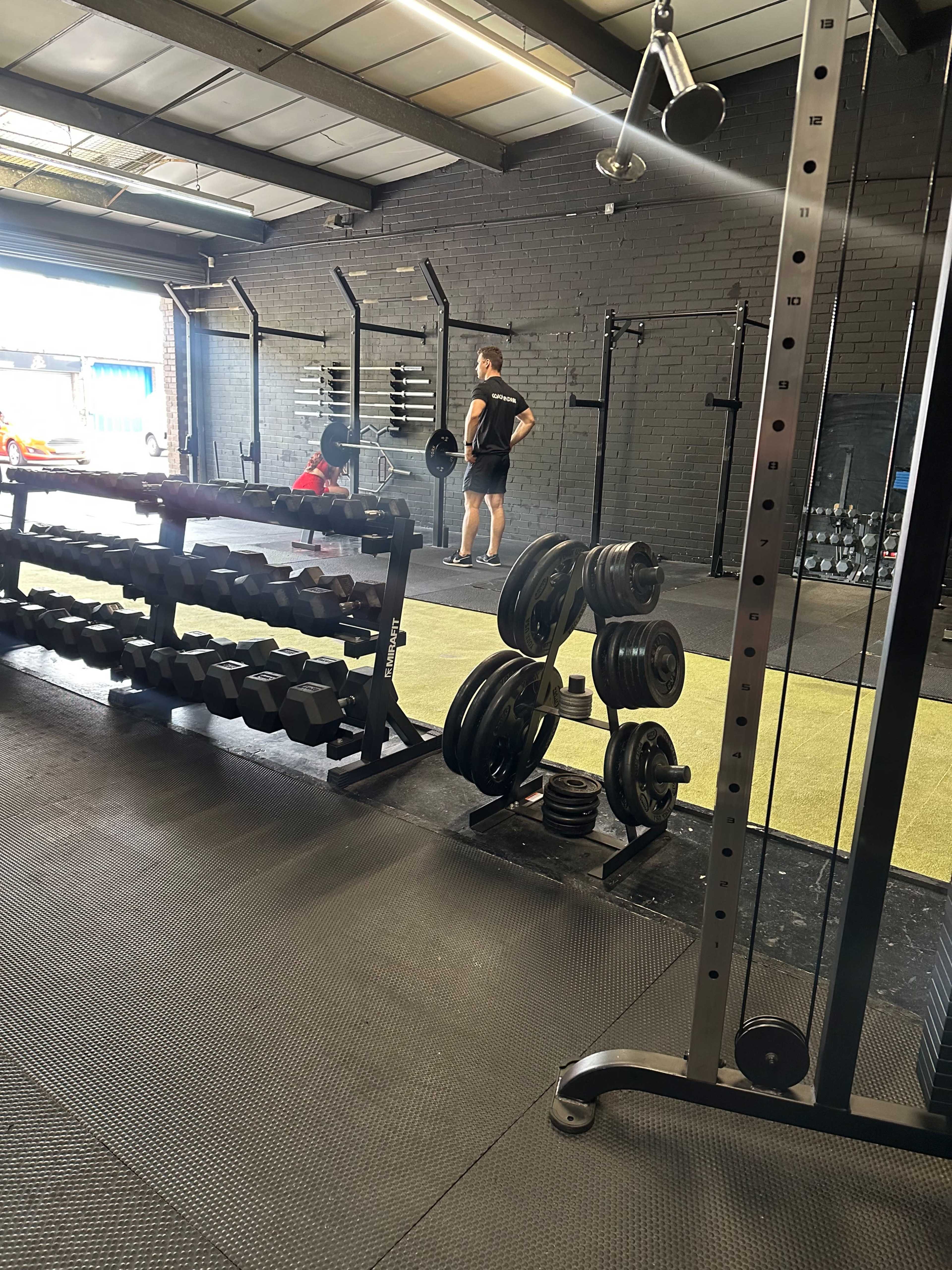 A person stands in a gym, facing a barbell, with various weights and equipment visible in the foreground.