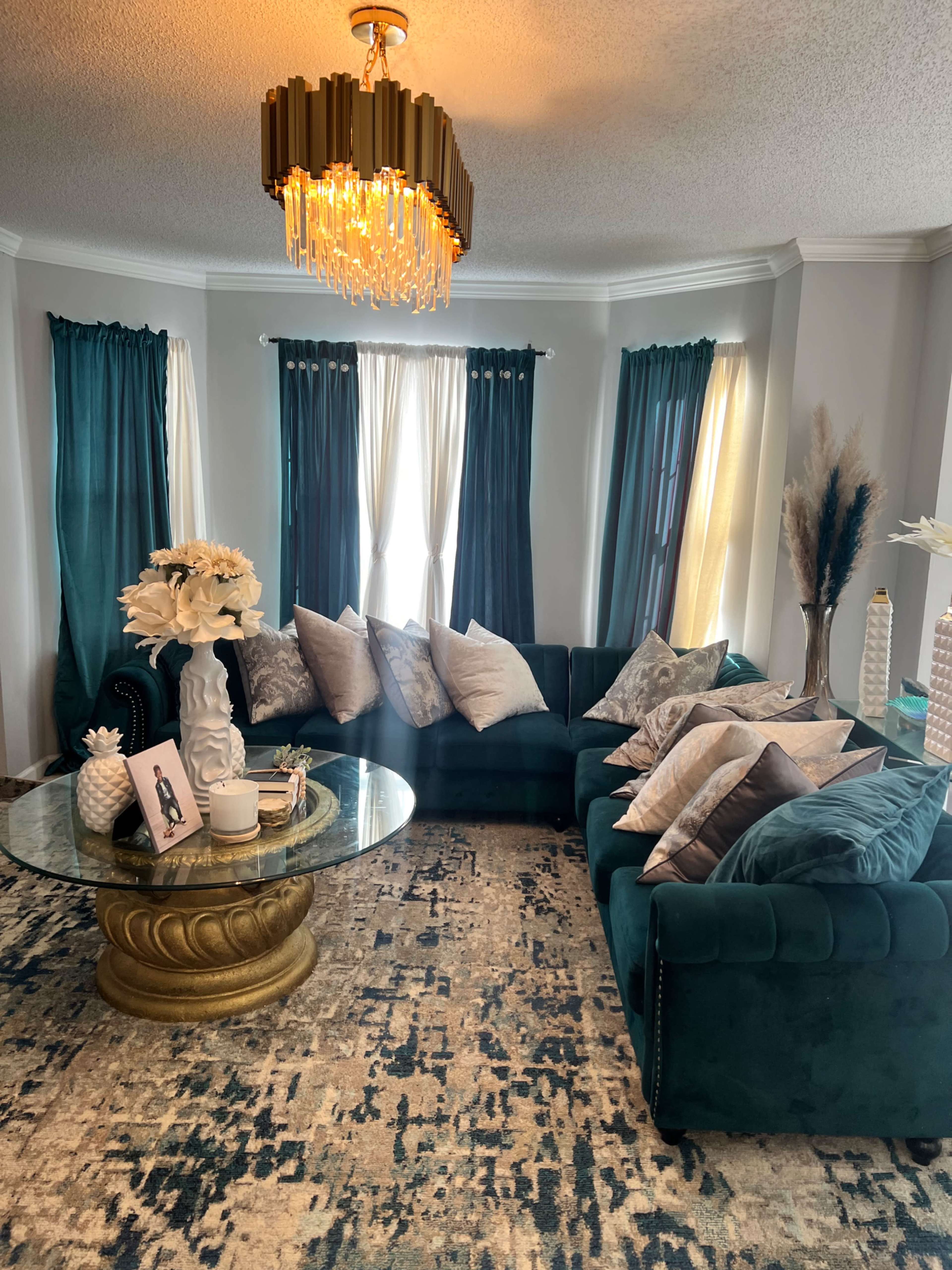 A cozy living room features a dark green sectional sofa, a round glass coffee table with decorative items, and large windows with sheer and solid curtains.