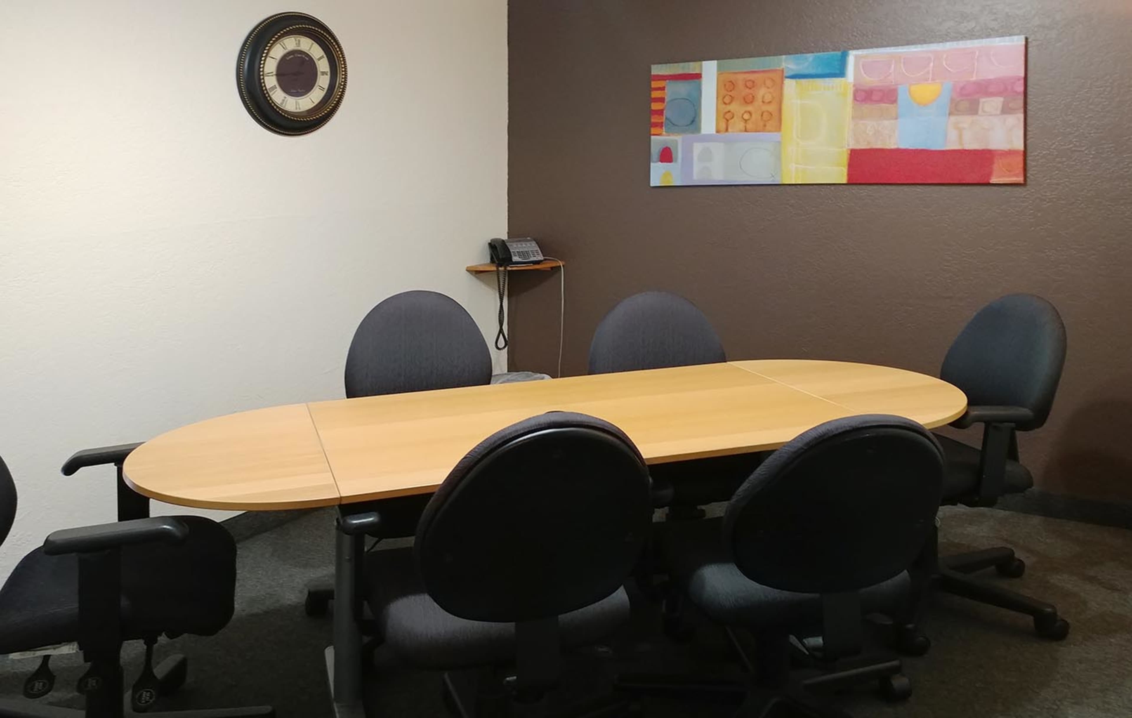A conference room features an oval wooden table surrounded by six rolling chairs, with a wall-mounted phone and abstract artwork in the background.