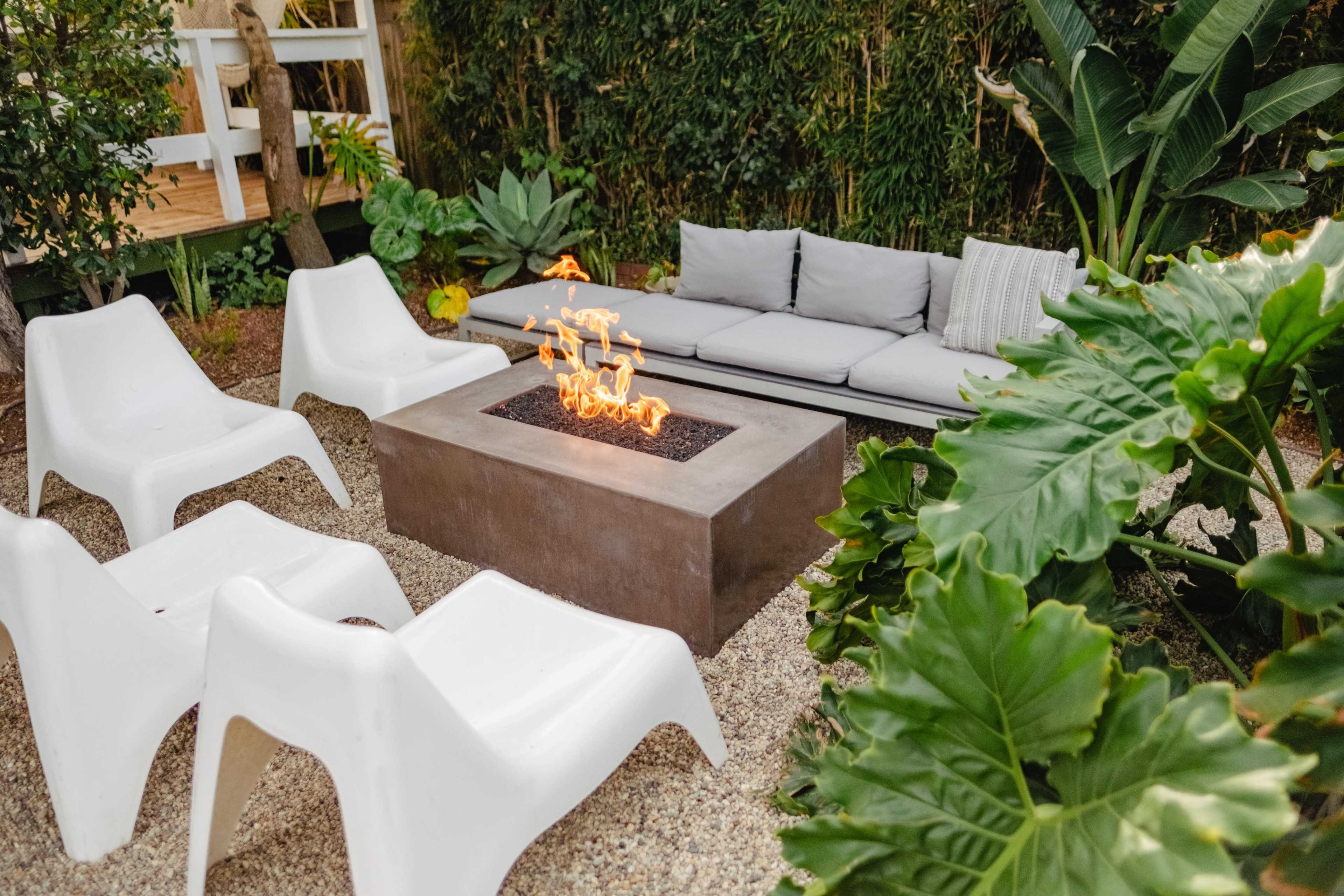 A modern outdoor seating area features a concrete fire pit surrounded by four white chairs and a gray couch, all set among lush greenery.
