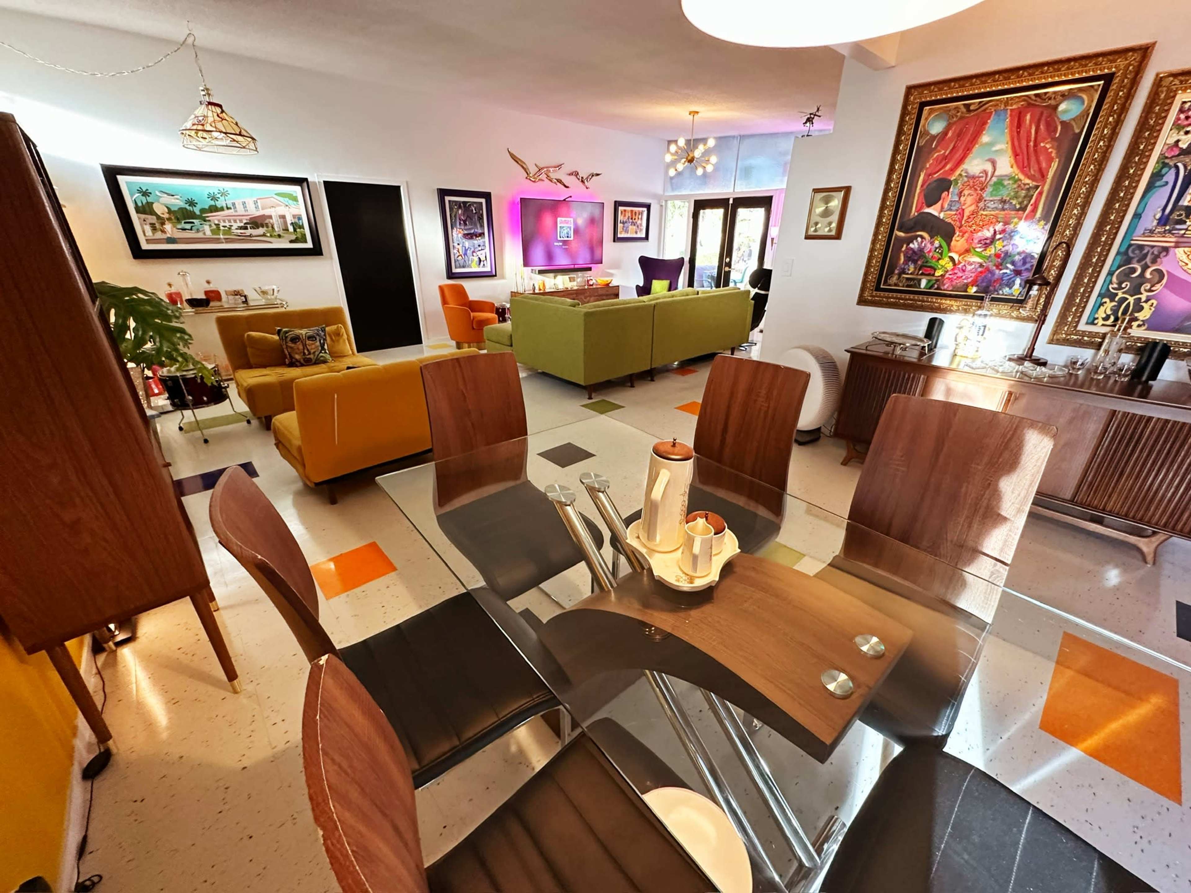 The image shows a brightly lit, modern living room and dining area with colorful furniture and artistic wall decorations.