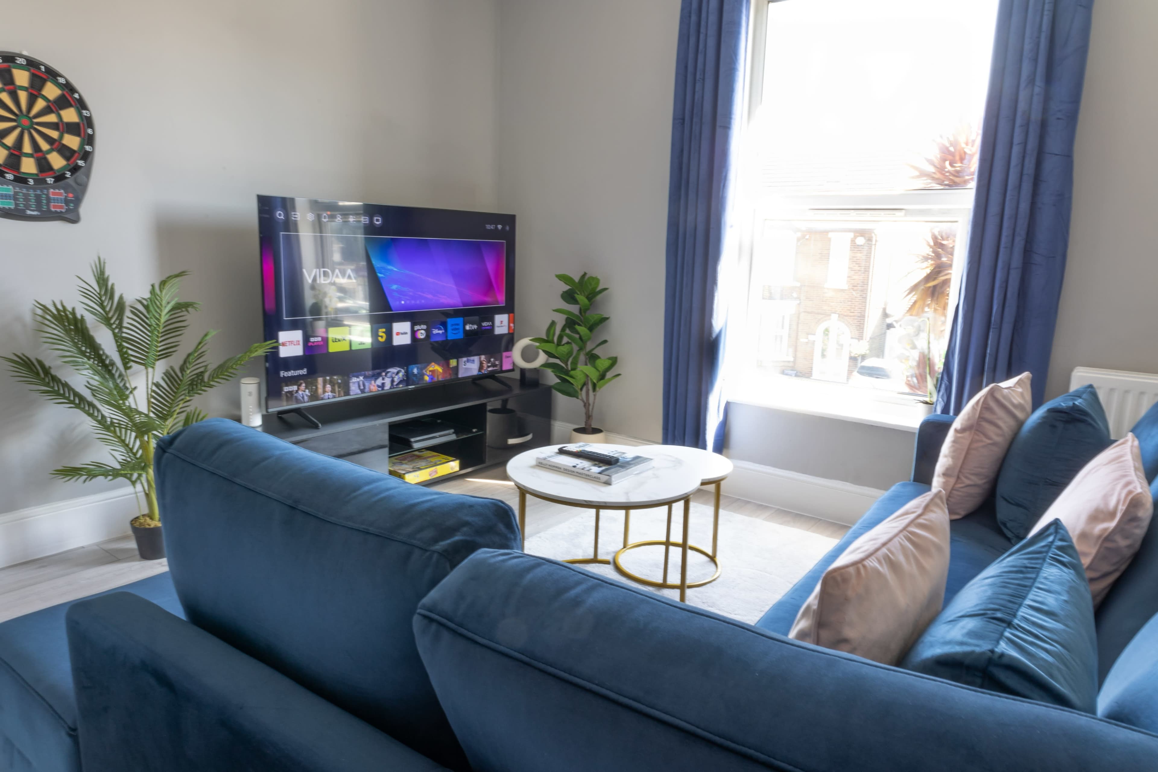 A living room with a blue sofa, a round coffee table, a television displaying a menu, and a potted plant near a window.