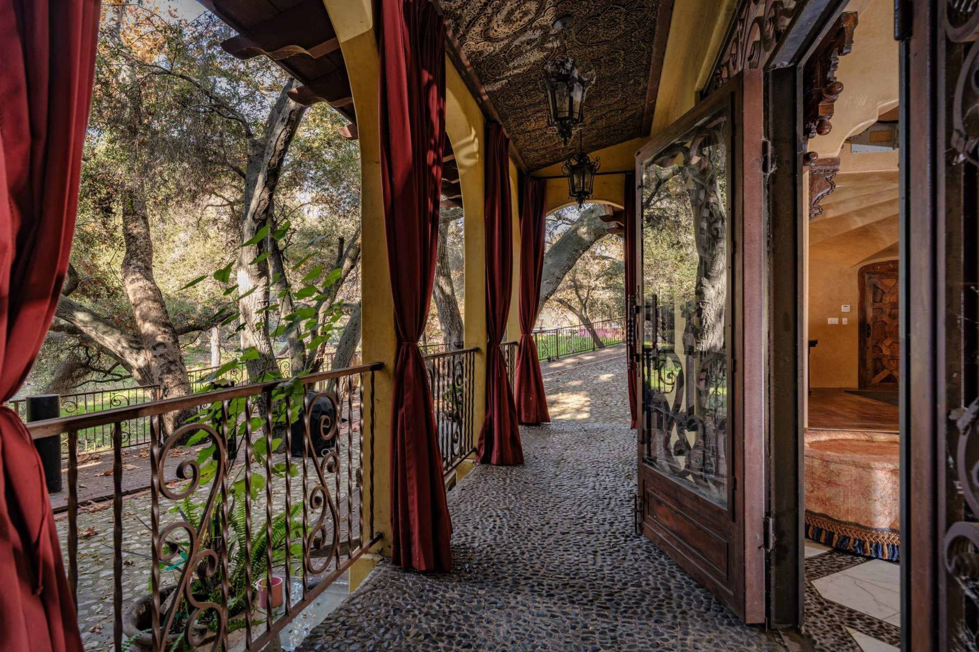 The image shows a balcony with a stone floor and decorative iron railings, framed by flowing red curtains and offering a view of a lush outdoor area.