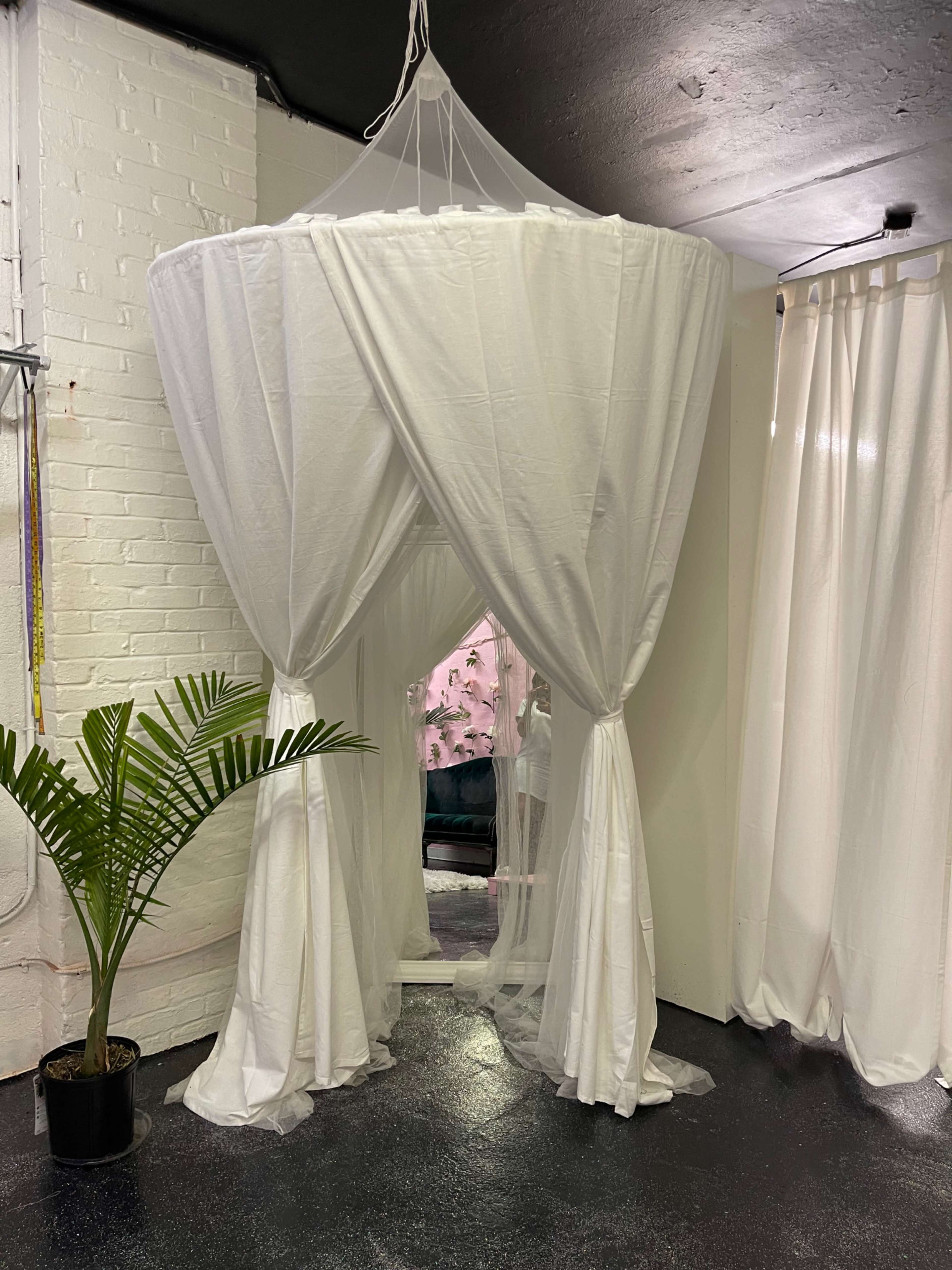 A large, white fabric canopy draped over a circular structure is situated in a room with white walls and a potted plant nearby.