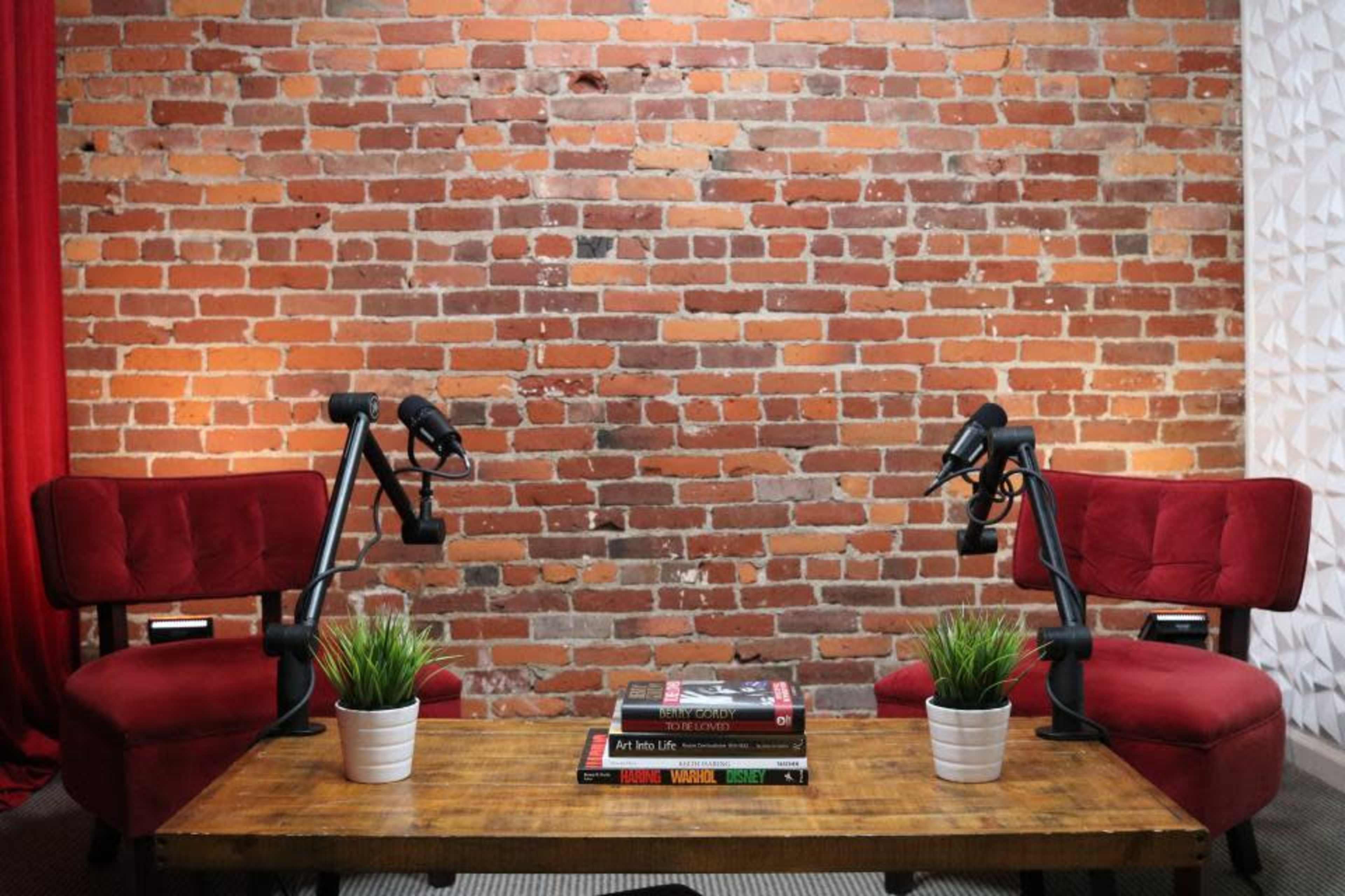 A small podcast studio featuring two red chairs with microphones on adjustable arms opposite a wooden table decorated with books and green plants against a brick wall.