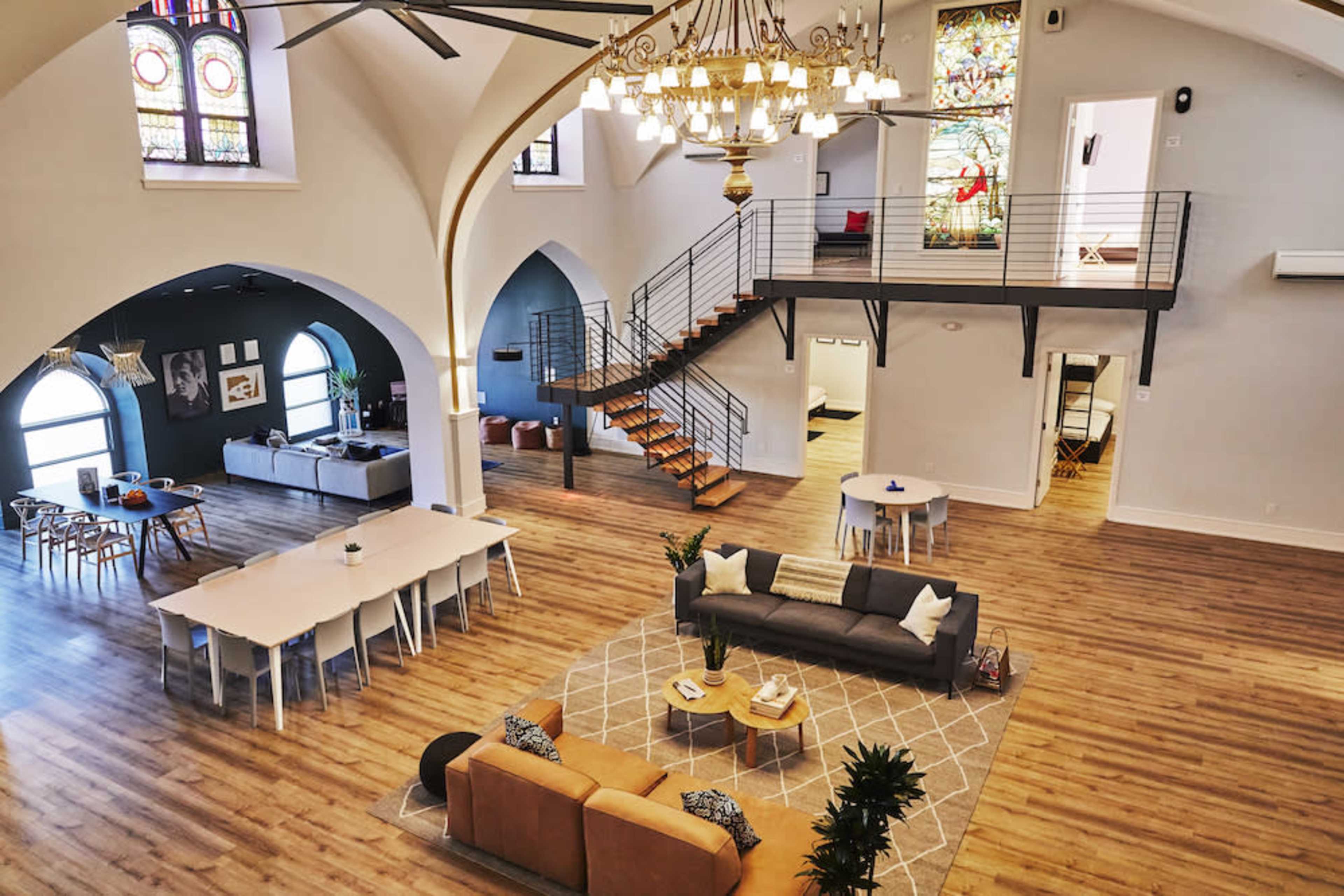 The image depicts a spacious interior of a renovated building featuring high ceilings, a chandelier, a mix of seating areas, and an elegant staircase leading to a second level.