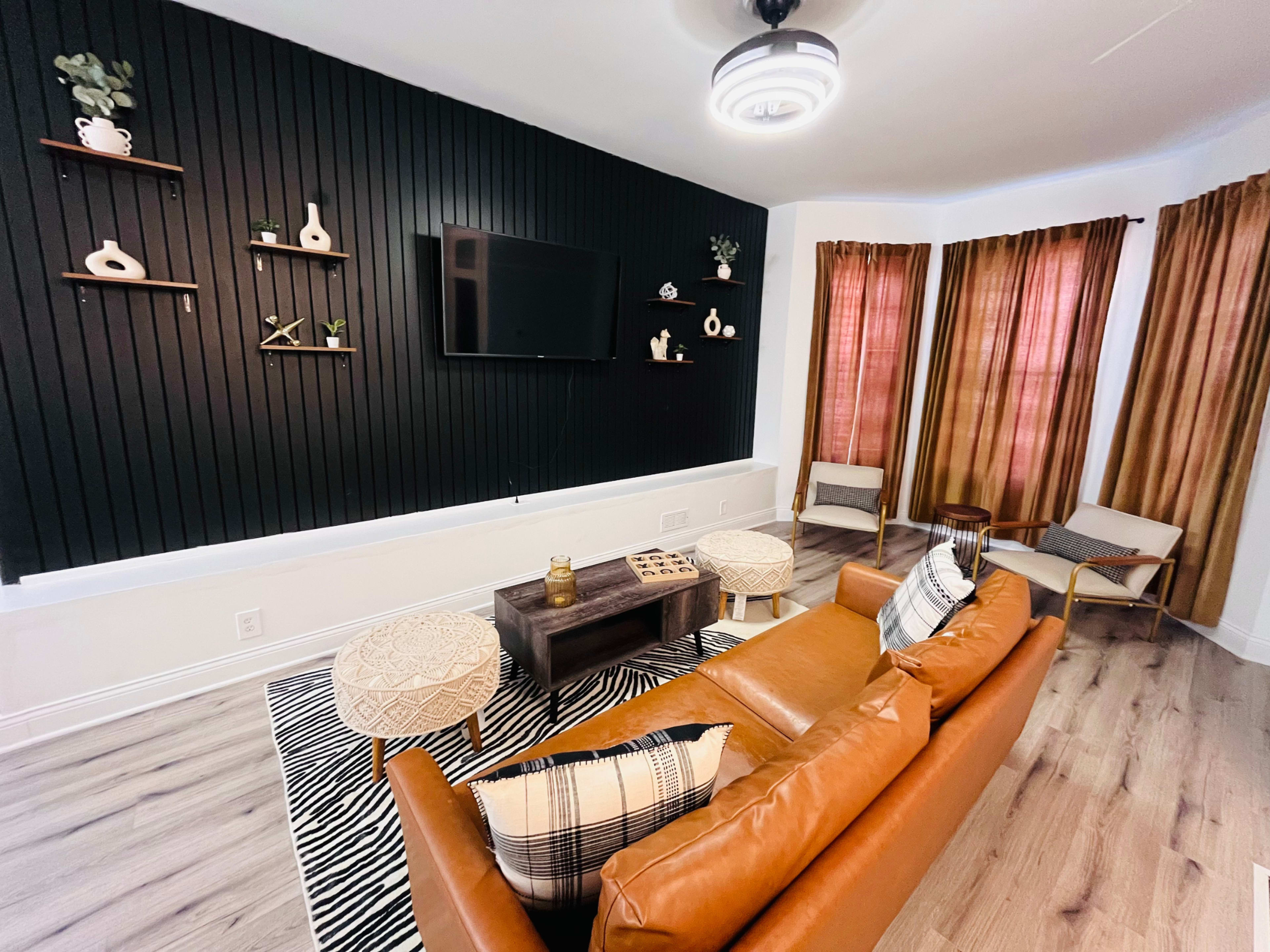 A modern living room featuring a brown leather sofa, decorative wall shelves, a television, and large windows with brown curtains.