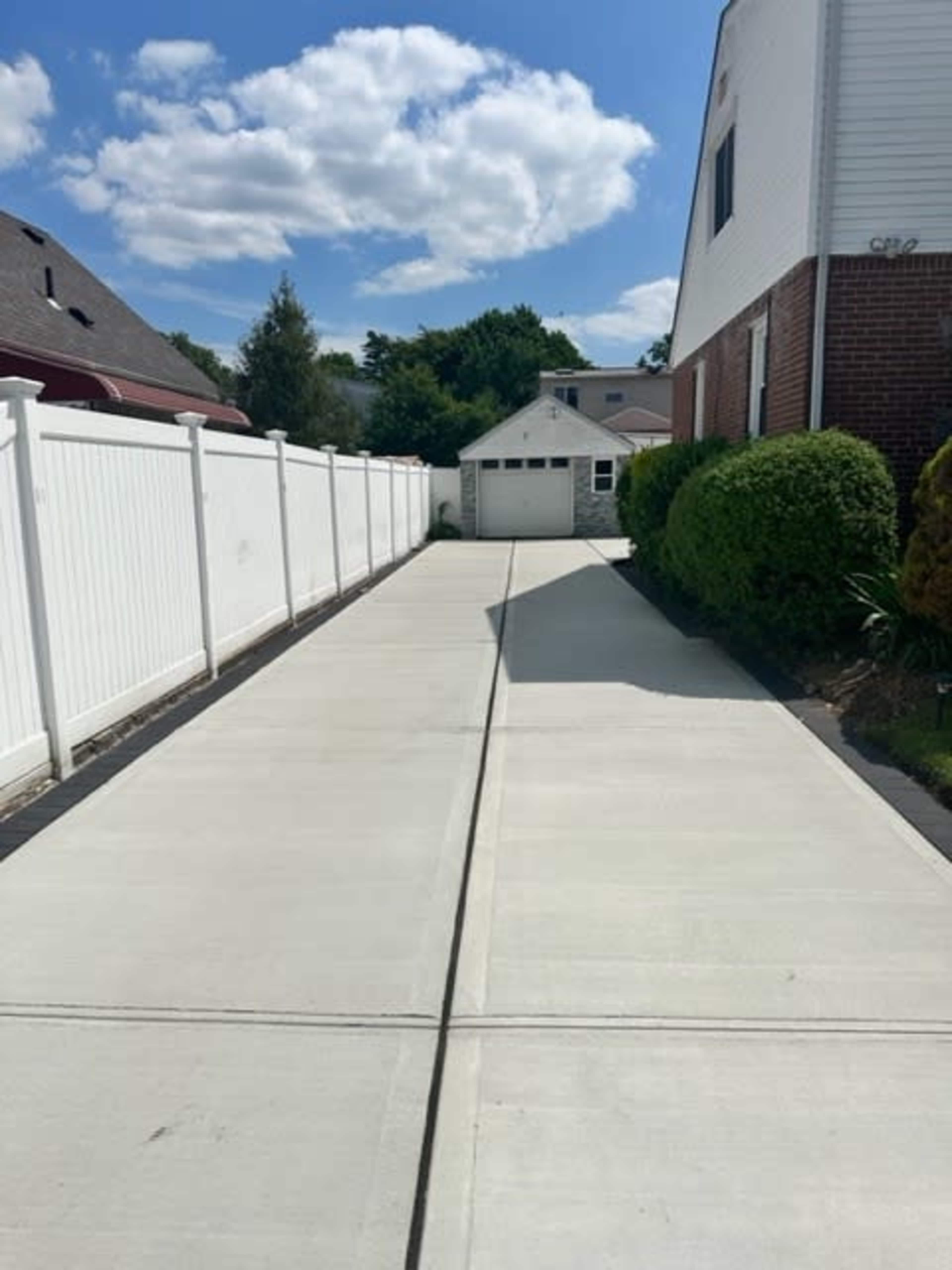 A concrete driveway leads to a garage at the end, bordered by a white fence and greenery on both sides.